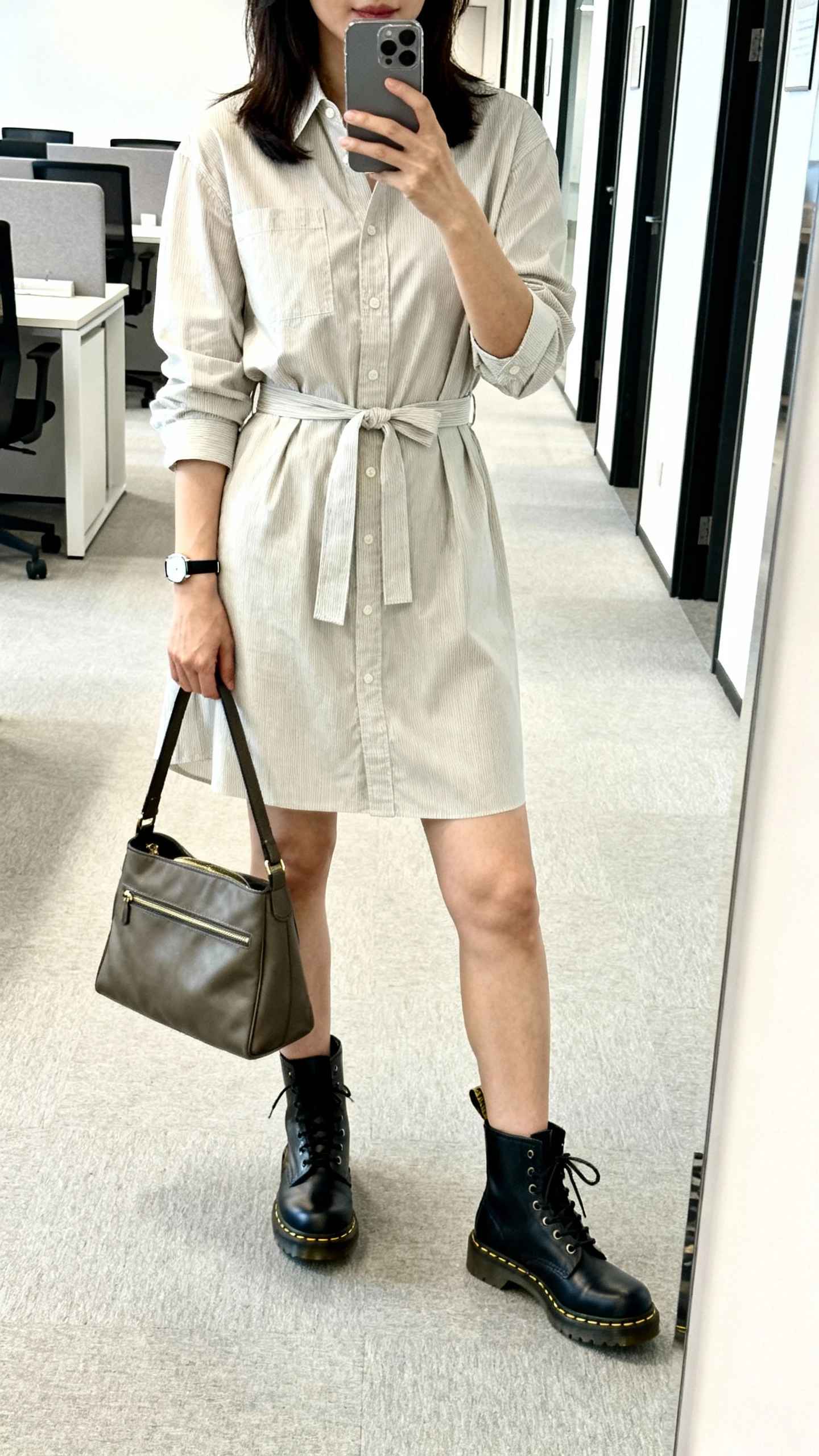 iPhone mirror selfie of a woman wearing a belted shirt dress with classic combat-style Doc Martens, structured tote and a slim watch, face not visible, cropped at shoulders, office hallway mirror, bright indoor lighting, iPhone photo quality.