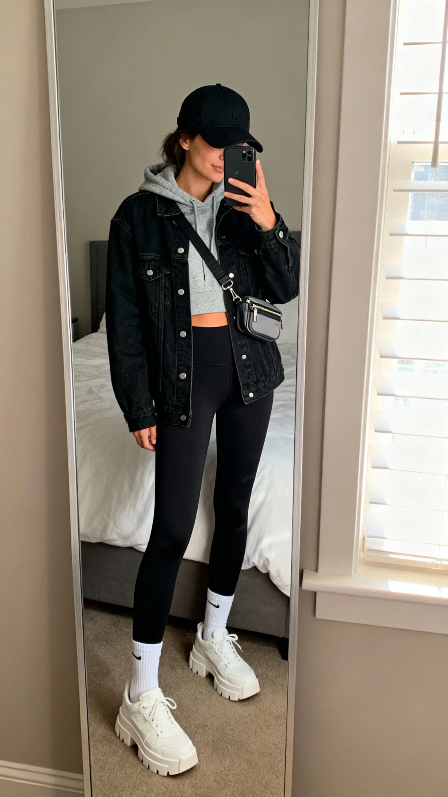 iPhone mirror selfie of a woman wearing a black denim jacket over a gray cropped hoodie, black high-waisted leggings, white crew socks, chunky white sneakers, a black baseball cap, and a small crossbody belt bag, face not visible, cropped at neck, bedroom mirror with morning window light, iPhone photo quality.