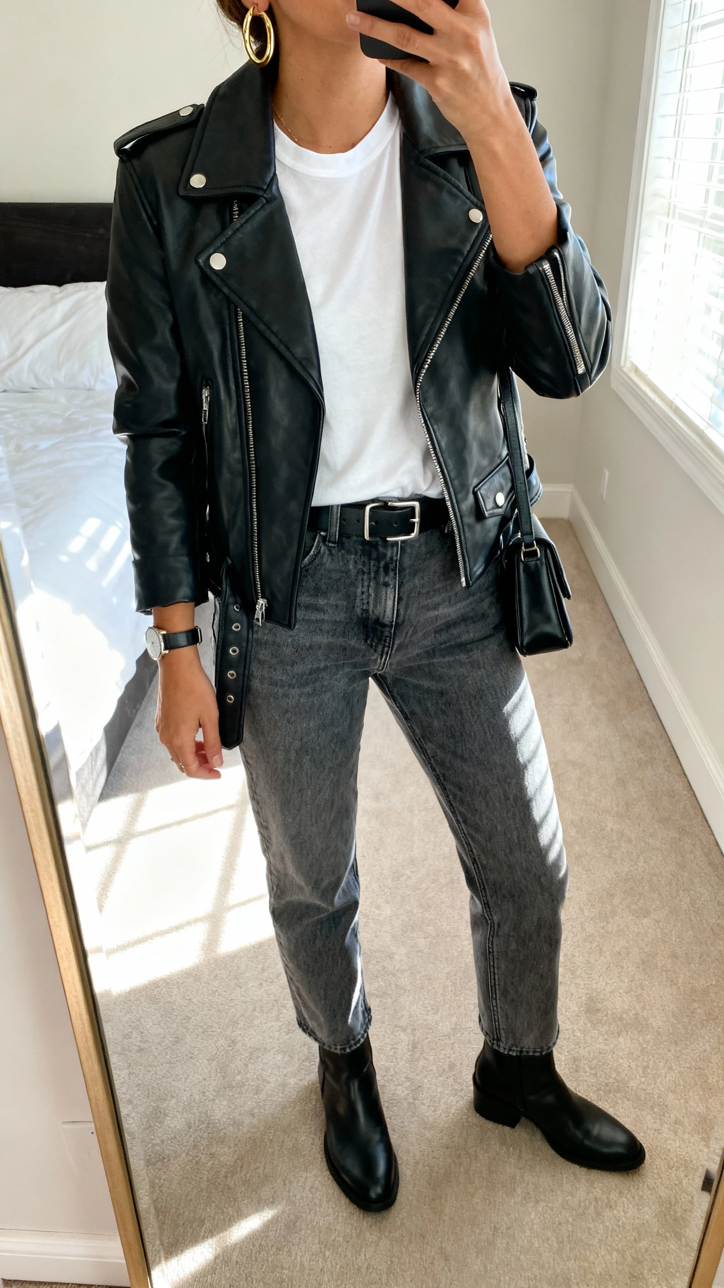 iPhone mirror selfie of a woman wearing a black leather moto jacket (hip-length), crisp white crewneck tee, mid-wash straight-leg jeans, black ankle boots, black belt with subtle metal buckle, gold hoop earrings, slim watch, and a structured black crossbody; face not visible, cropped at neck, bedroom mirror with morning window light, iPhone photo quality.
