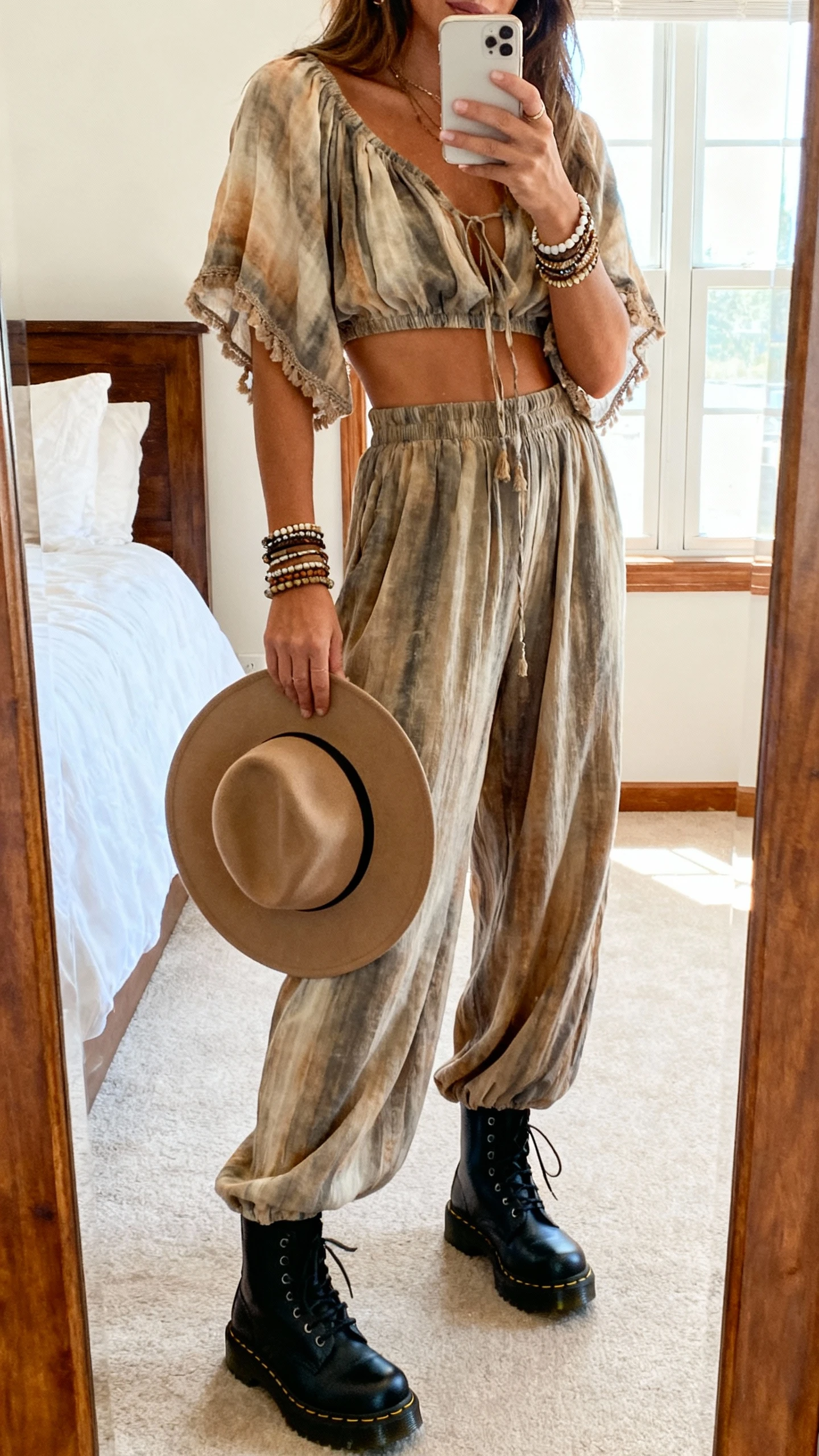 iPhone mirror selfie of a woman wearing a boho matching set (flowy crop top and wide-leg pants) with chunky black Doc Martens, layered bracelets, and a floppy hat in hand, face not visible, bedroom full-length mirror, natural window light, iPhone photo quality.