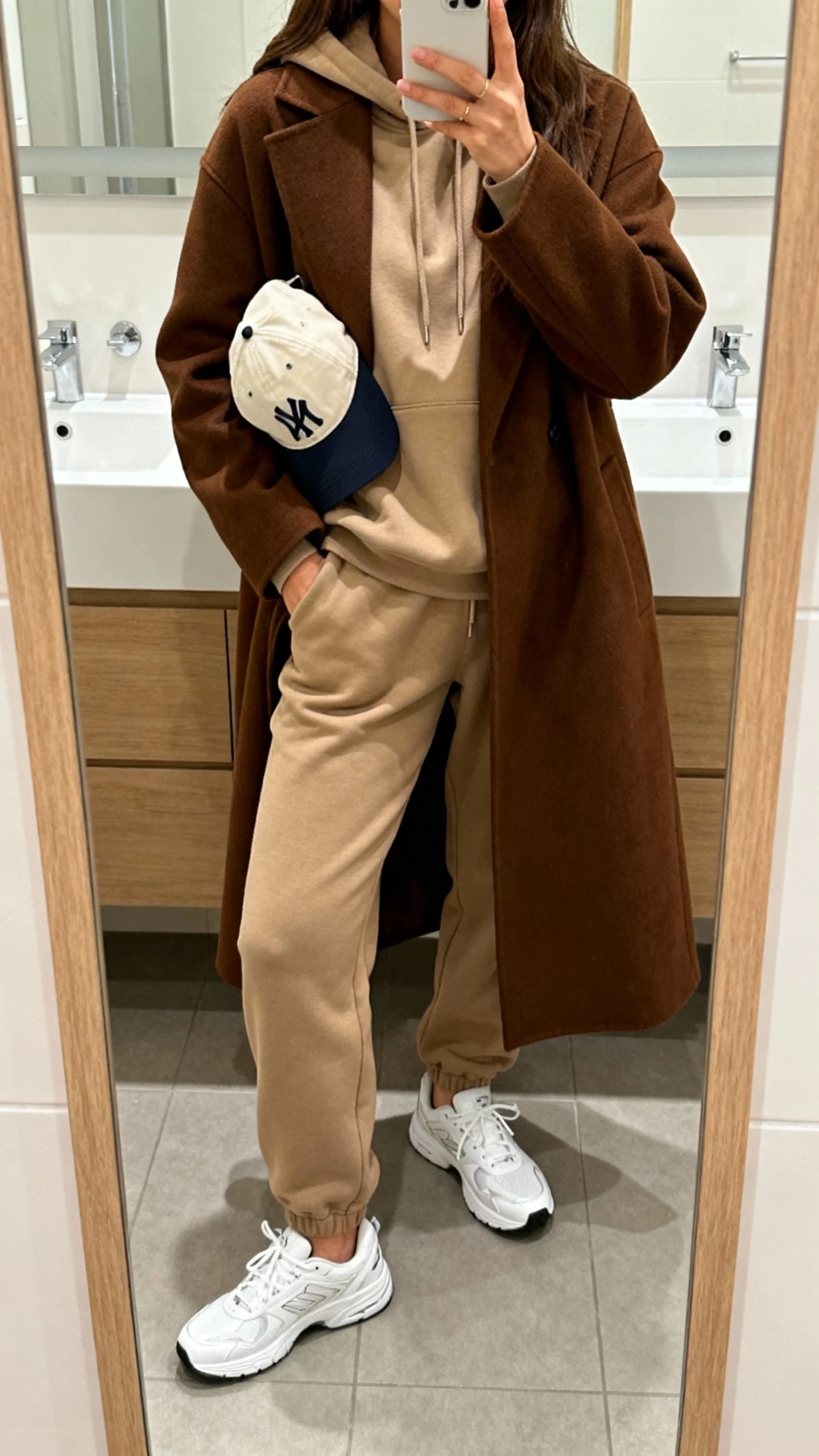 iPhone mirror selfie of a woman wearing a brown coat over a sand-toned hoodie, matching joggers, clean white sneakers, and a baseball cap tucked under her arm, face not visible, bathroom mirror, soft indoor lighting, iPhone photo quality.