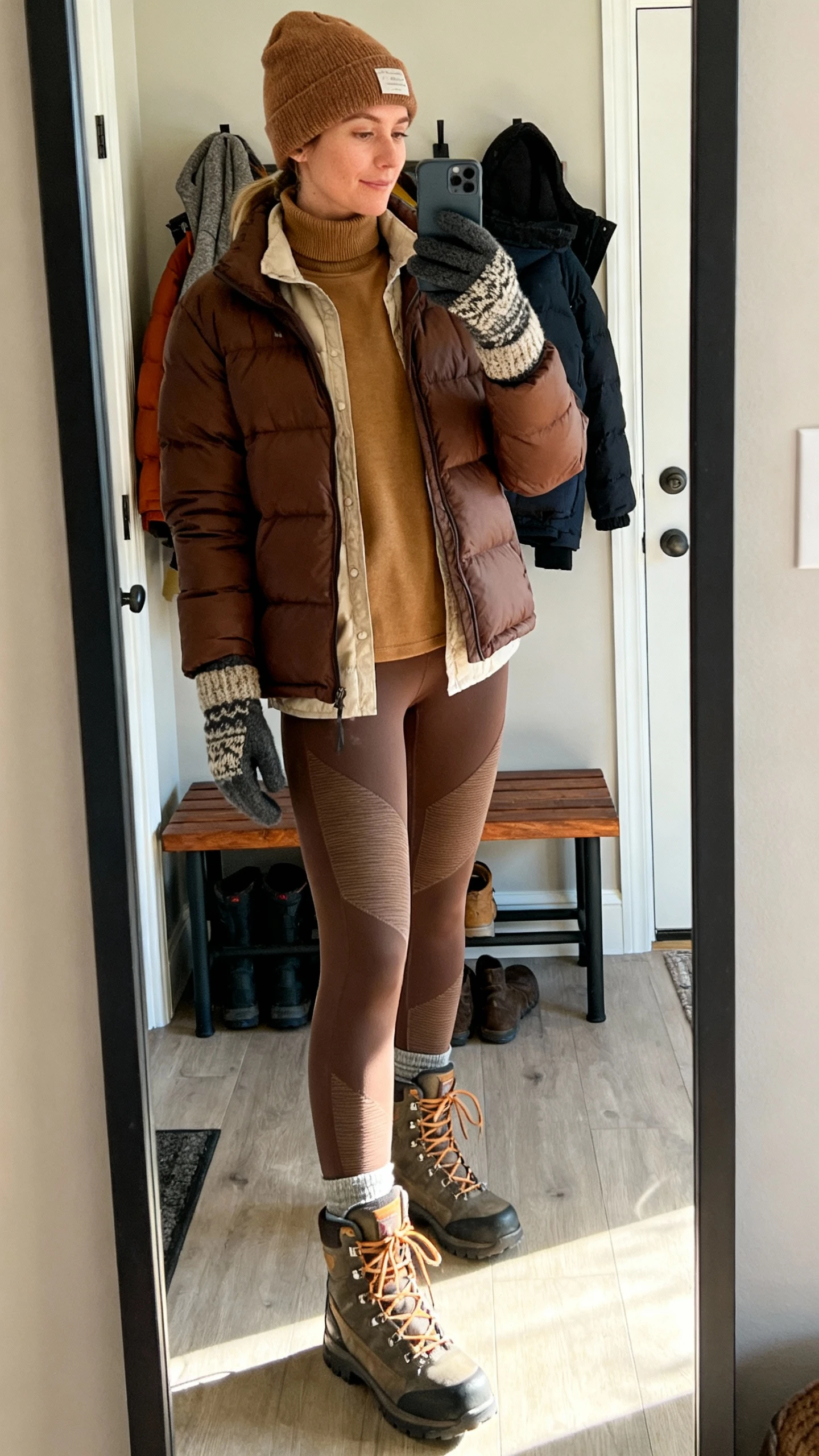 iPhone mirror selfie of a woman wearing a brown puffer layered over a tan turtleneck, thermal leggings, chunky lace-up boots, beanie, and knit gloves, face not visible, cropped at neck, entryway mirror with winter gear bench, cool morning daylight, iPhone photo quality.