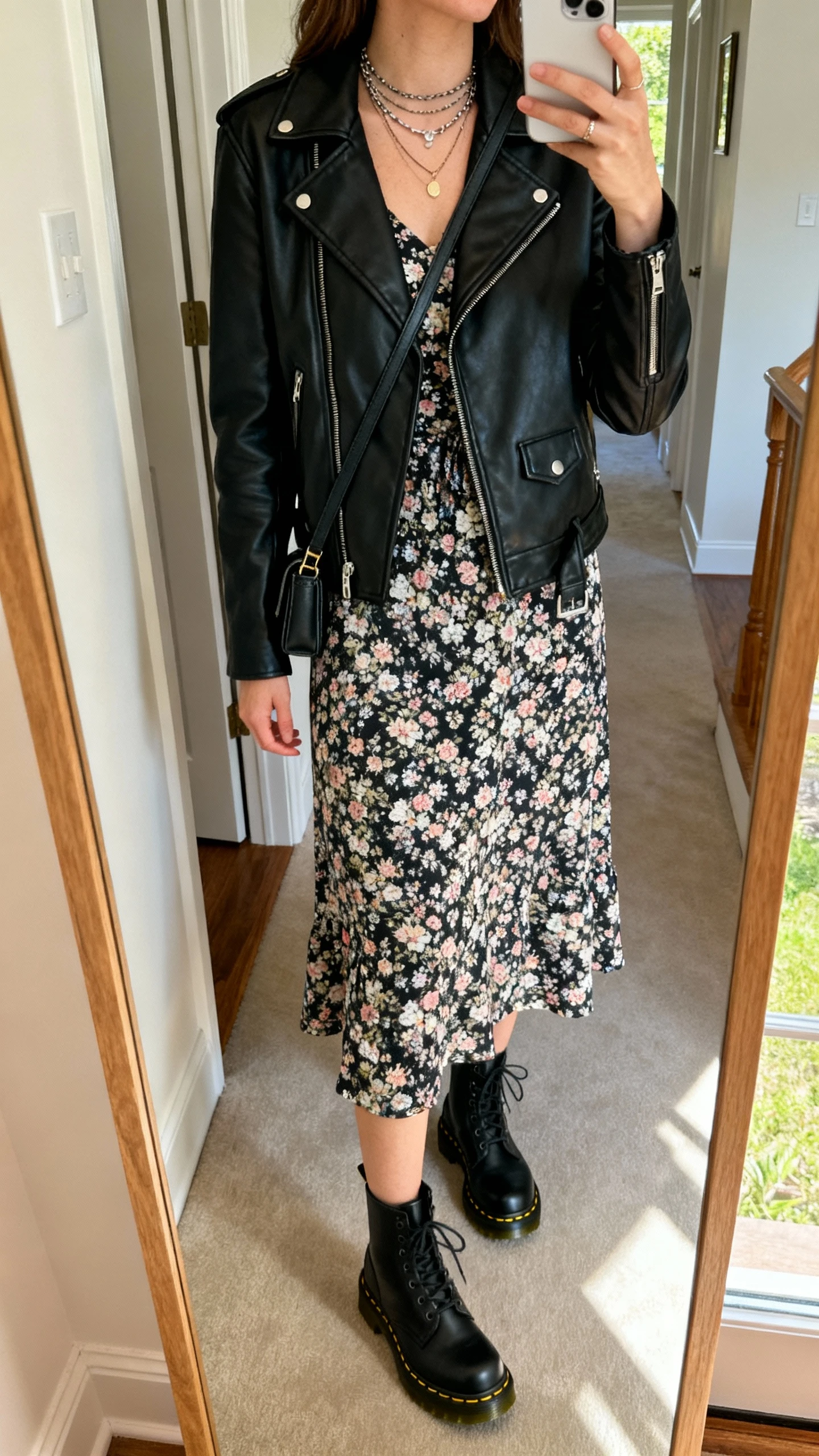 iPhone mirror selfie of a woman wearing a casual floral midi dress topped with a black leather biker jacket, black Doc Martens boots, layered necklaces, and a small crossbody, face not visible, cropped at neck, full-length mirror in entryway, afternoon indoor light, iPhone photo quality.