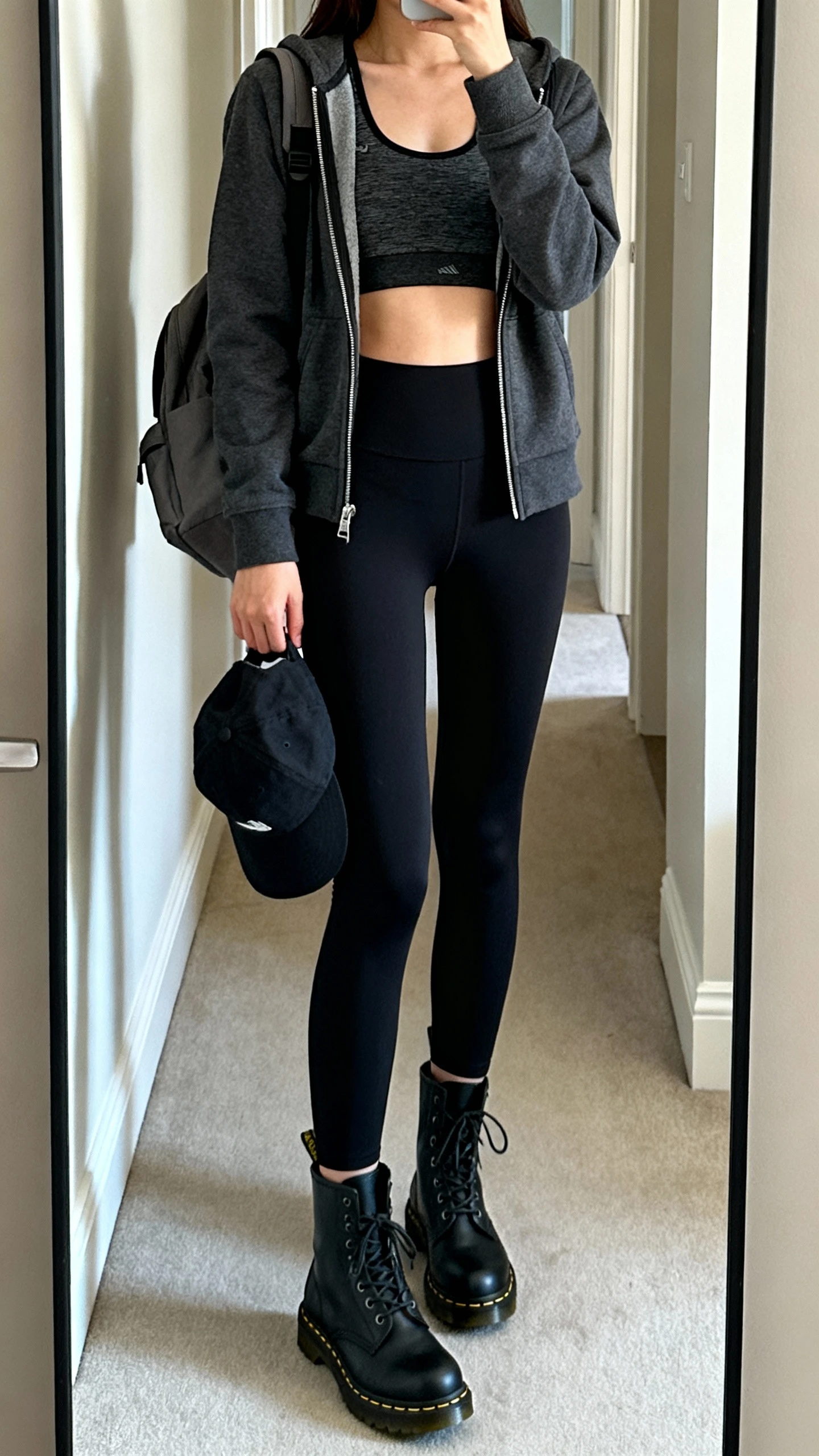 iPhone mirror selfie of a woman wearing a charcoal zip-up hoodie layered over a cropped athletic tank with high-waisted black leggings, black Doc Martens boots, baseball cap in hand, and a minimal backpack, face not visible, cropped at shoulders, hallway full-length mirror, indoor daylight, iPhone photo quality.