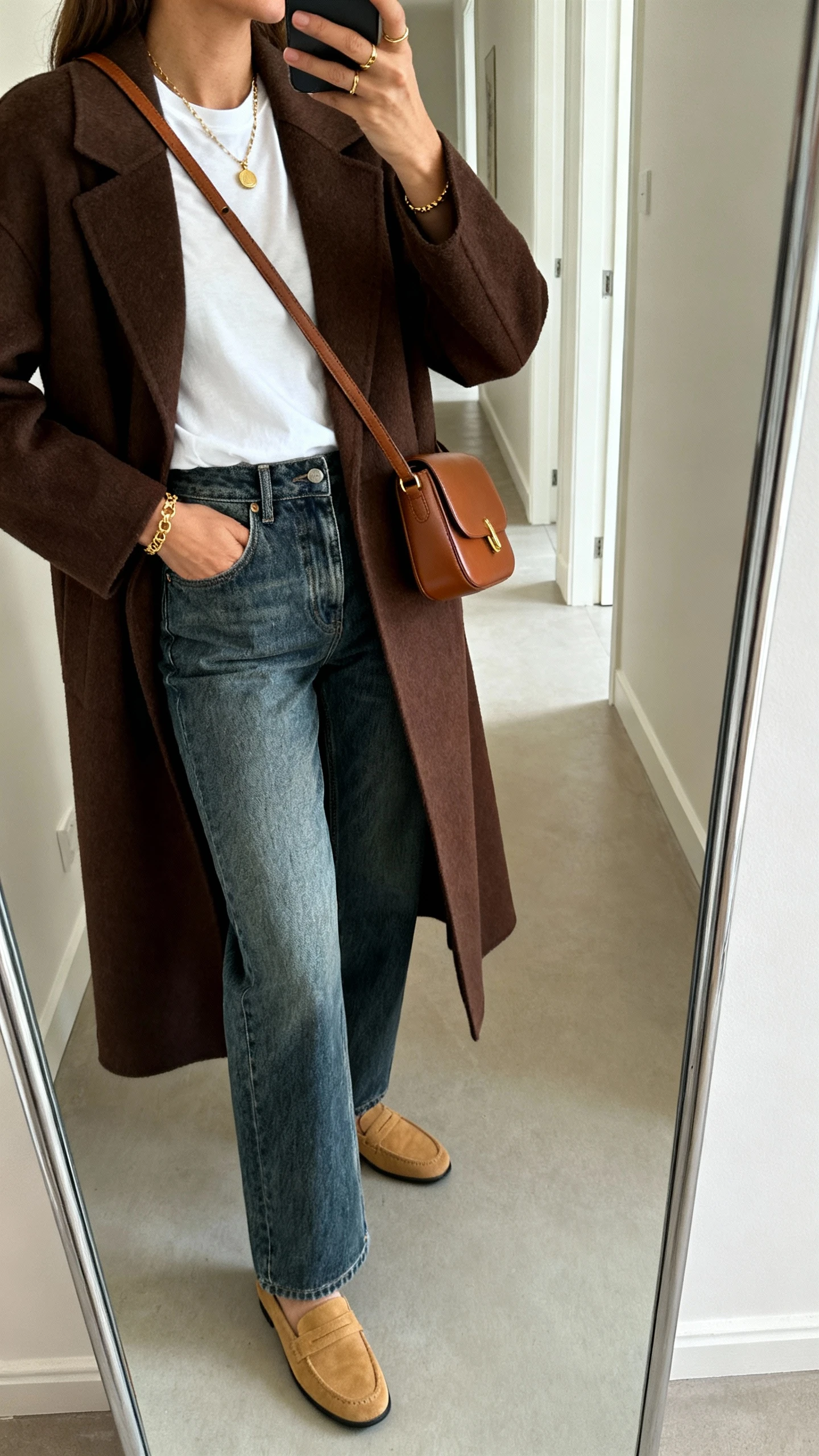 iPhone mirror selfie of a woman wearing a chocolate brown coat layered over a crisp white tee, high-rise medium-wash straight-leg jeans, tan loafers, and a brown crossbody bag with subtle gold jewelry, face not visible, side angle in hallway mirror, natural indoor light, iPhone photo quality.