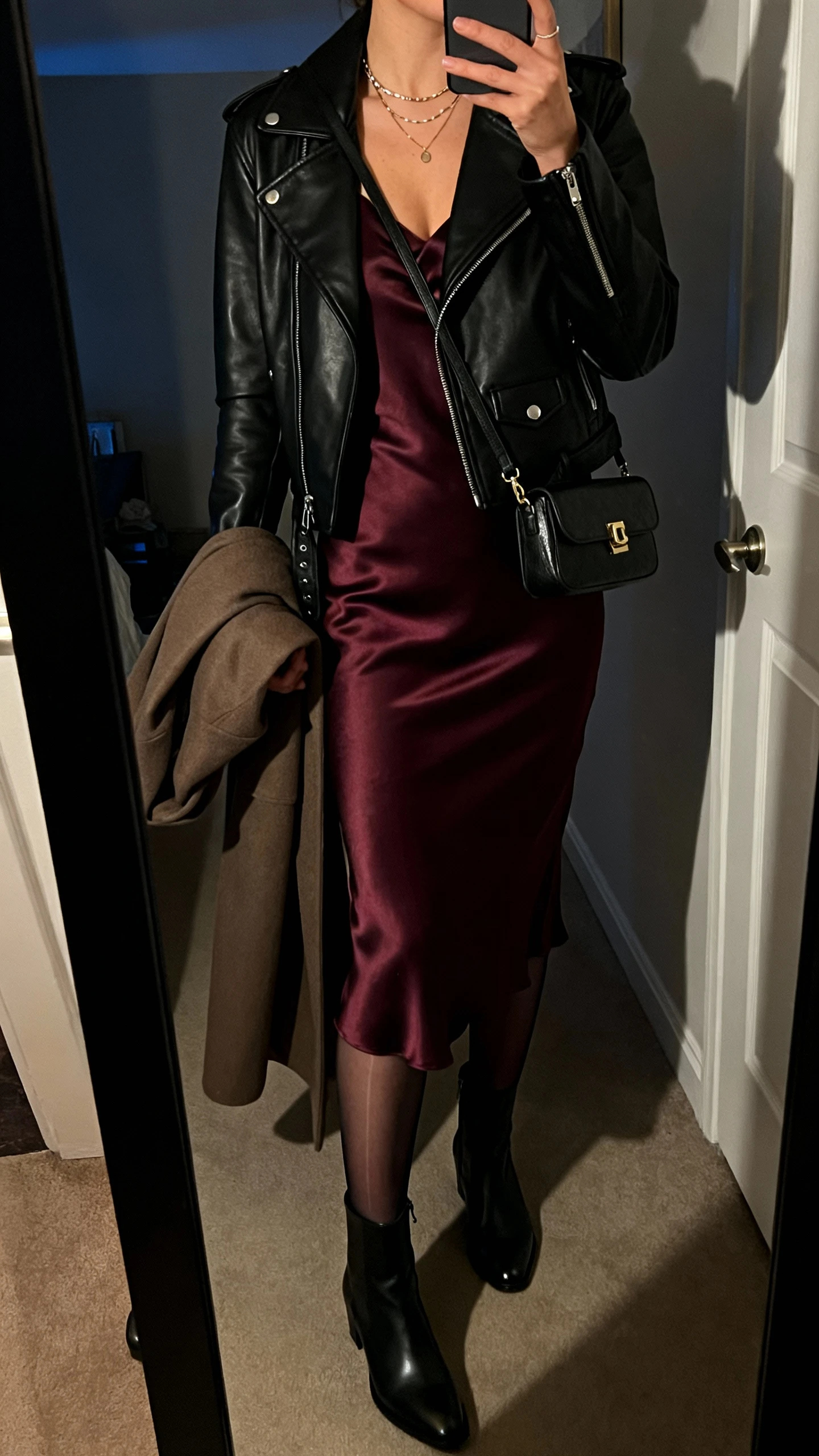 iPhone mirror selfie of a woman wearing a classic black leather moto over a bias-cut satin slip dress in deep wine, sleek ankle boots, layered delicate necklaces, and a small shoulder bag; face not visible, cropped at neck, dim indoor evening light, optional sheer tights and longline coat draped over arm, iPhone photo quality.