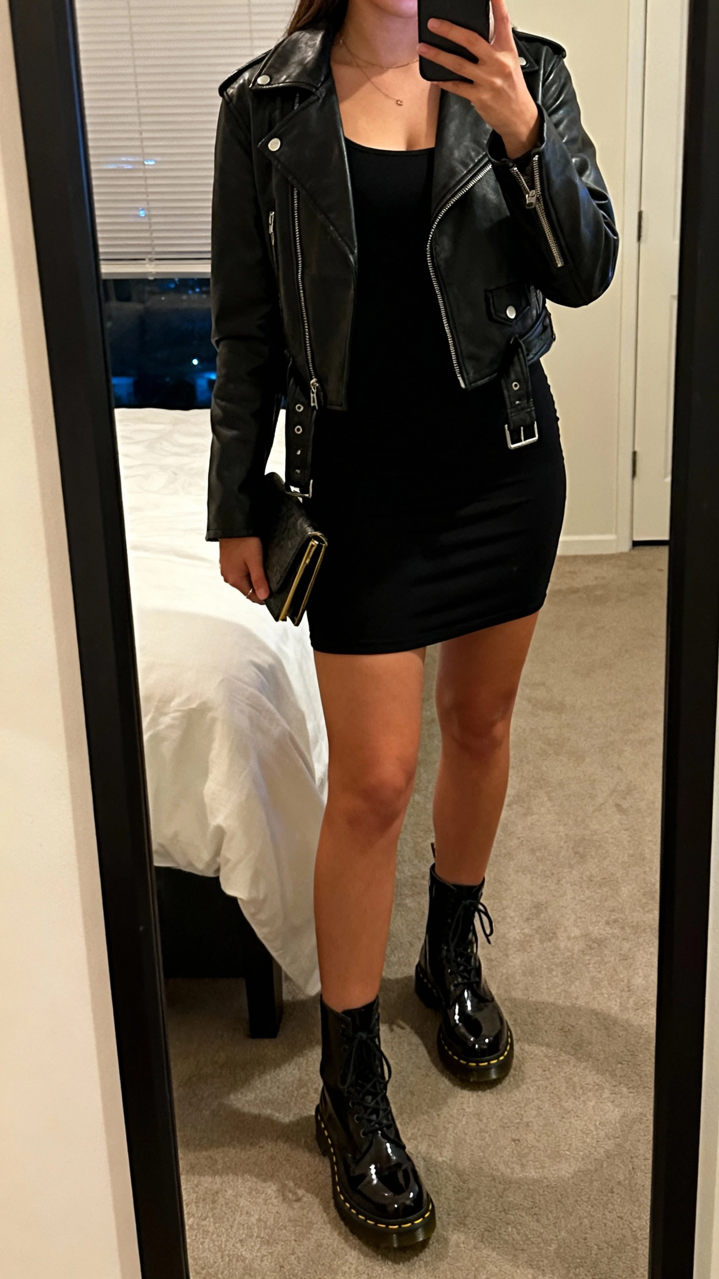 iPhone mirror selfie of a woman wearing a fitted little black dress with glossy patent Doc Martens boots, leather moto jacket and a small clutch, face not visible, cropped at neck, bedroom mirror, evening indoor lighting, iPhone photo quality.