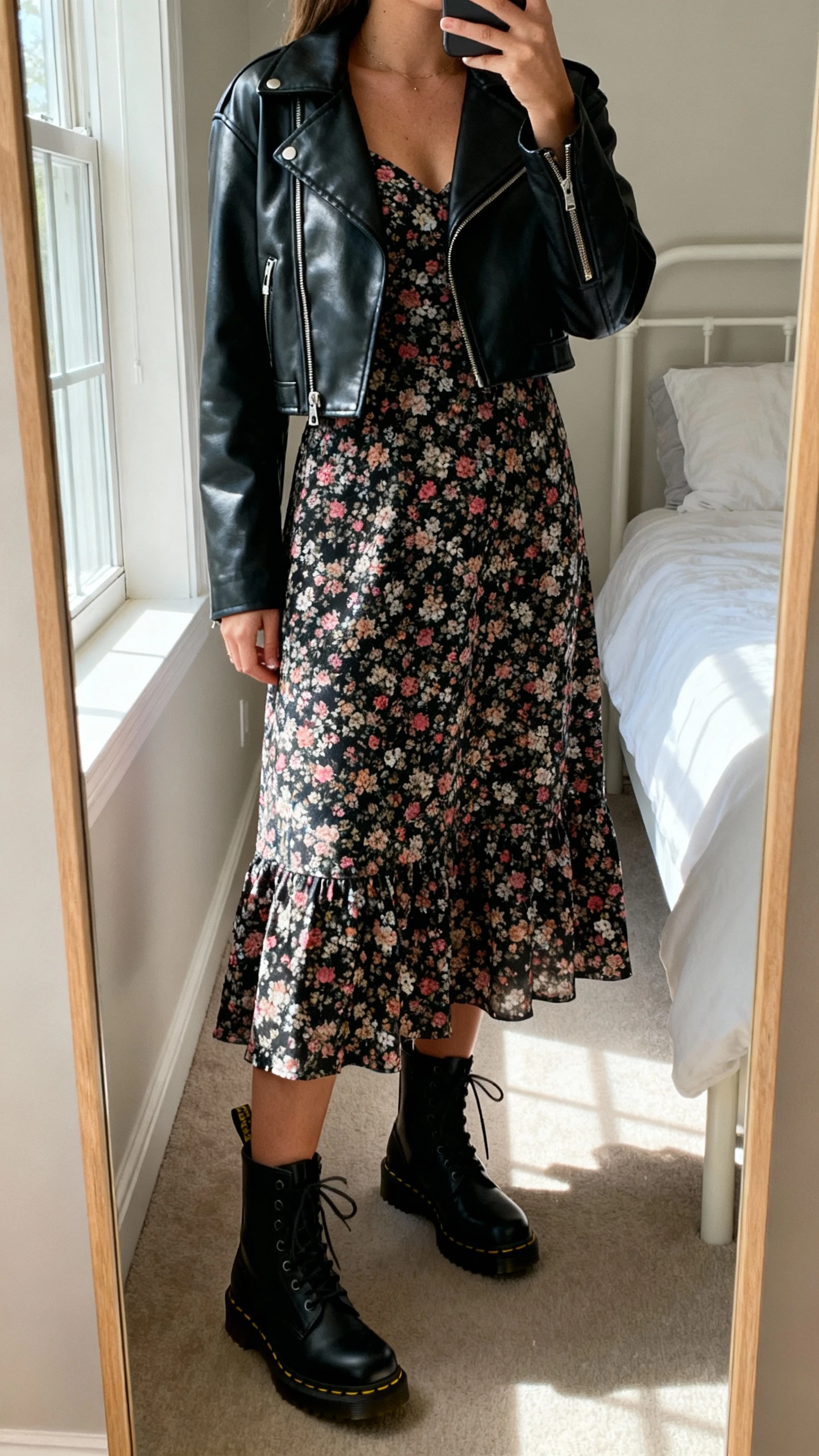 iPhone mirror selfie of a woman wearing a floral midi dress paired with black Doc Martens combat boots and a cropped leather jacket, face not visible, cropped at neck, bedroom mirror with natural window light, iPhone photo quality.