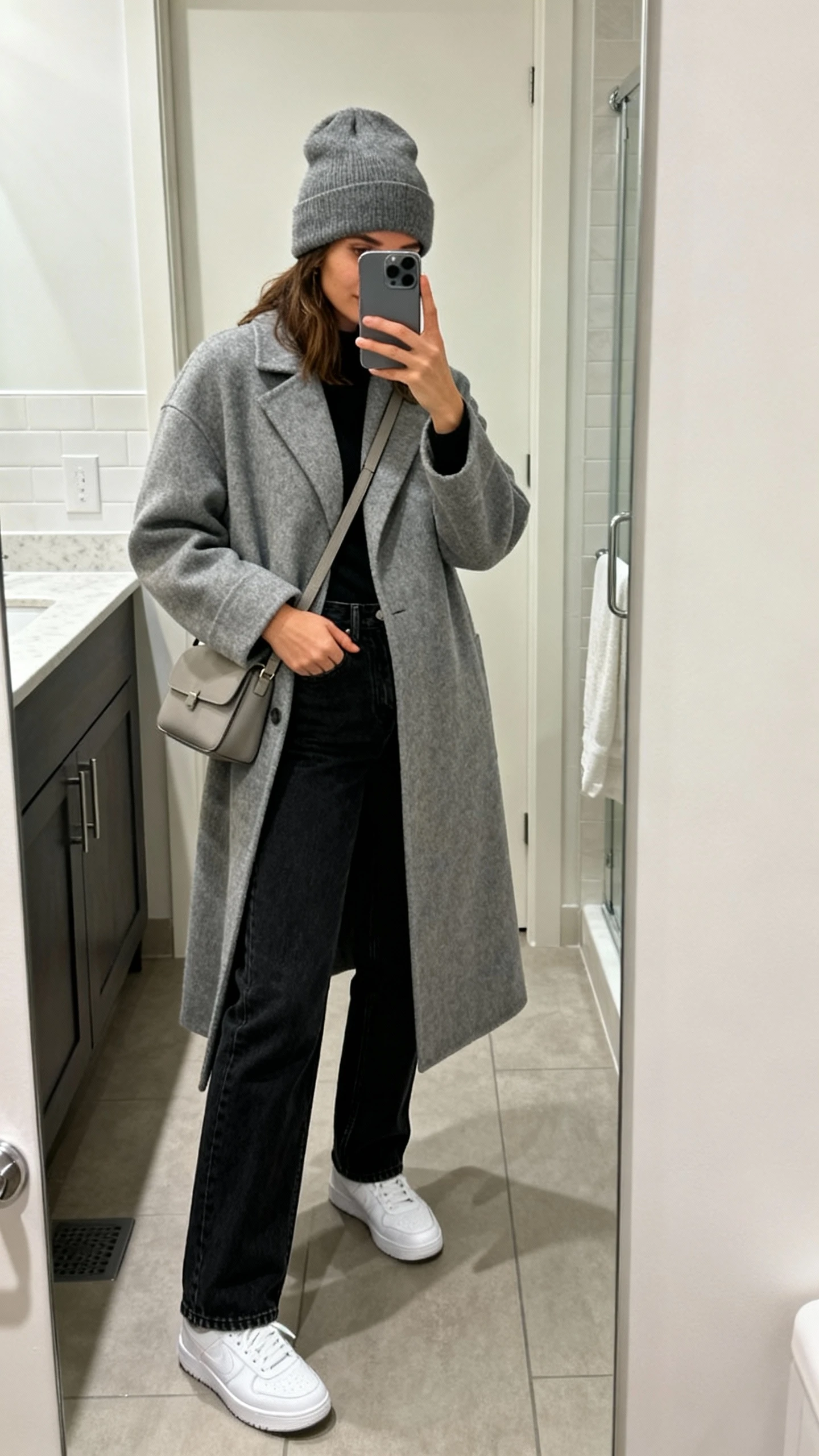 iPhone mirror selfie of a woman wearing a grey coat with white sneakers, black straight-leg jeans, a grey beanie, and a simple crossbody bag, face not visible, bathroom mirror, casual indoor lighting, iPhone photo quality