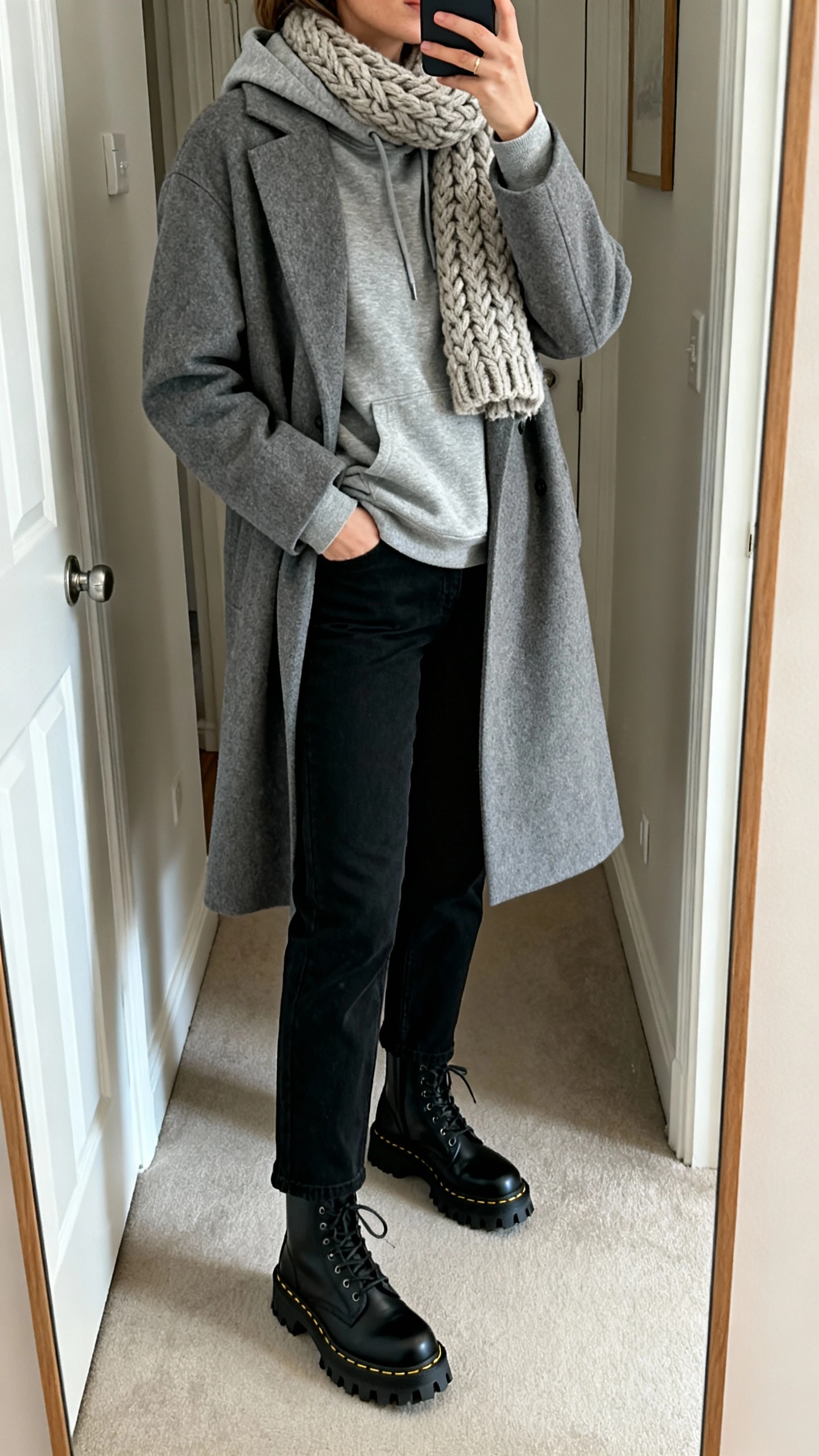 iPhone mirror selfie of a woman wearing a grey coat over a heather grey hoodie, chunky wool scarf, black straight-leg pants, and black lug-sole boots, face not visible, cropped at neck, entryway full-length mirror, soft indoor lighting, iPhone photo quality