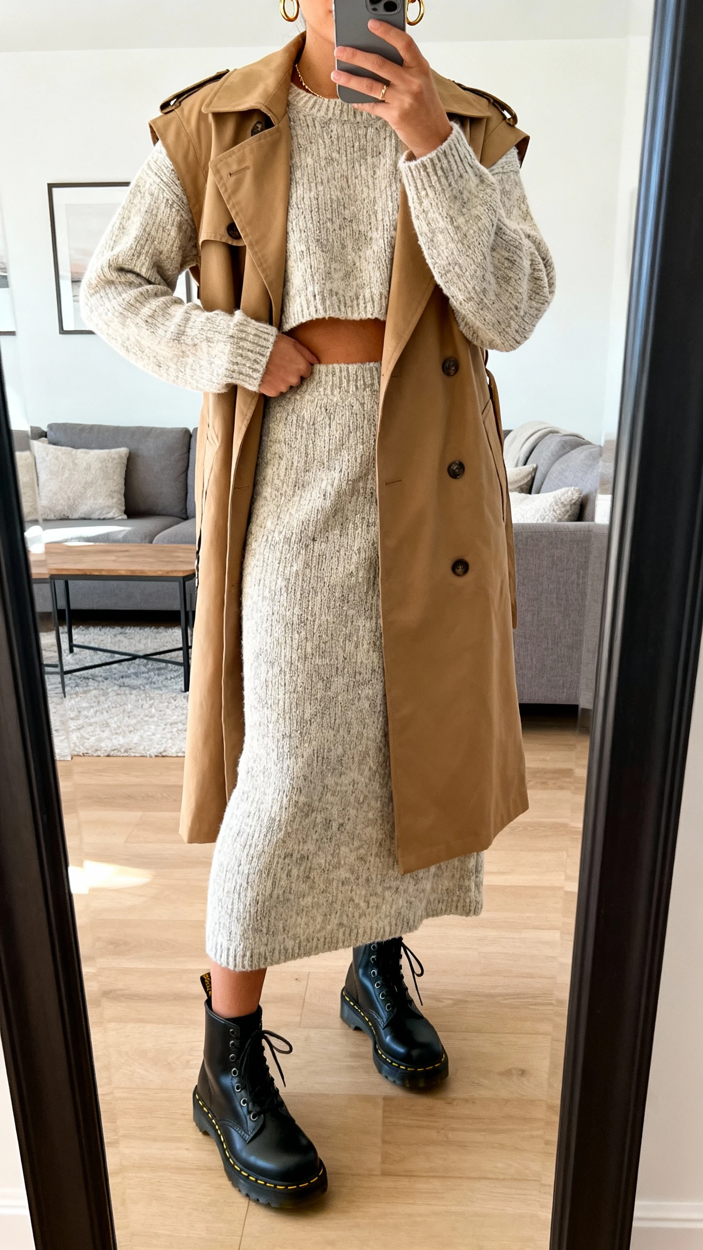 iPhone mirror selfie of a woman wearing a knit co-ord set (cropped sweater and matching midi skirt) layered with a tan trench vest, black Doc Martens, and delicate gold hoops, face not visible, cropped at neck, living room full-length mirror, soft morning light, iPhone photo quality.