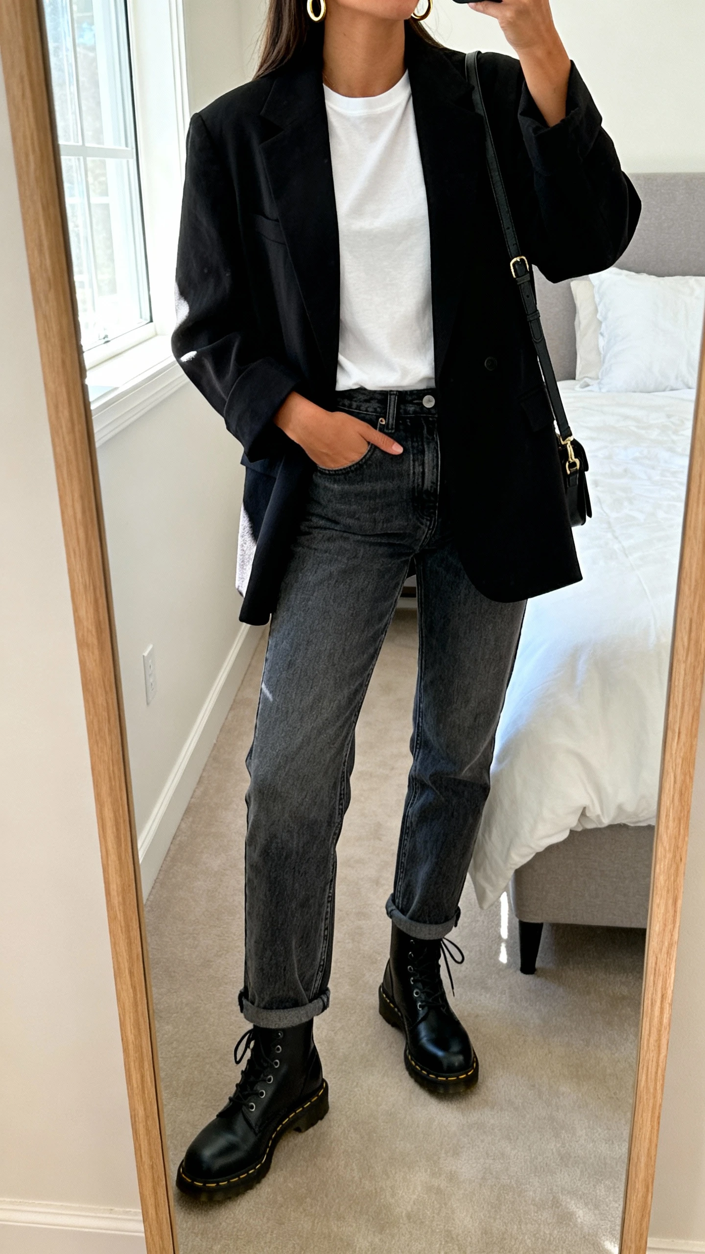 iPhone mirror selfie of a woman wearing a relaxed black blazer over a white tee, straight-leg mid-wash jeans, black Doc Martens boots, simple gold hoops, and a crossbody bag, face not visible, cropped at neck, bedroom mirror with natural window light, iPhone photo quality.