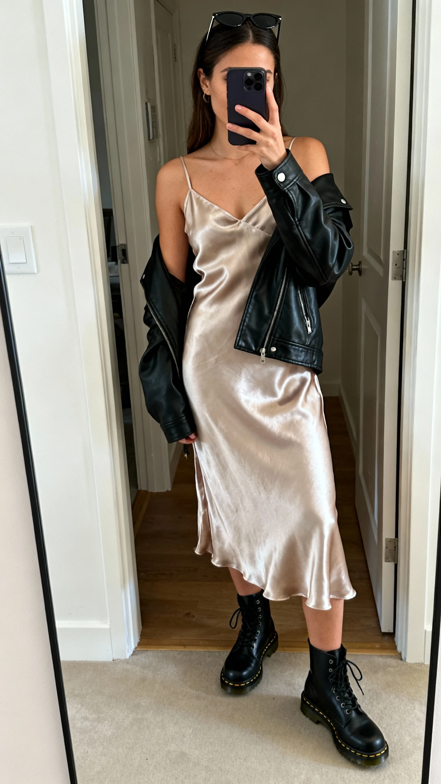 iPhone mirror selfie of a woman wearing a satin slip dress with a black leather jacket draped over shoulders, black Doc Martens, slim sunglasses perched on head, face not visible, cropped at shoulders, entryway mirror, indoor ambient light, iPhone photo quality.