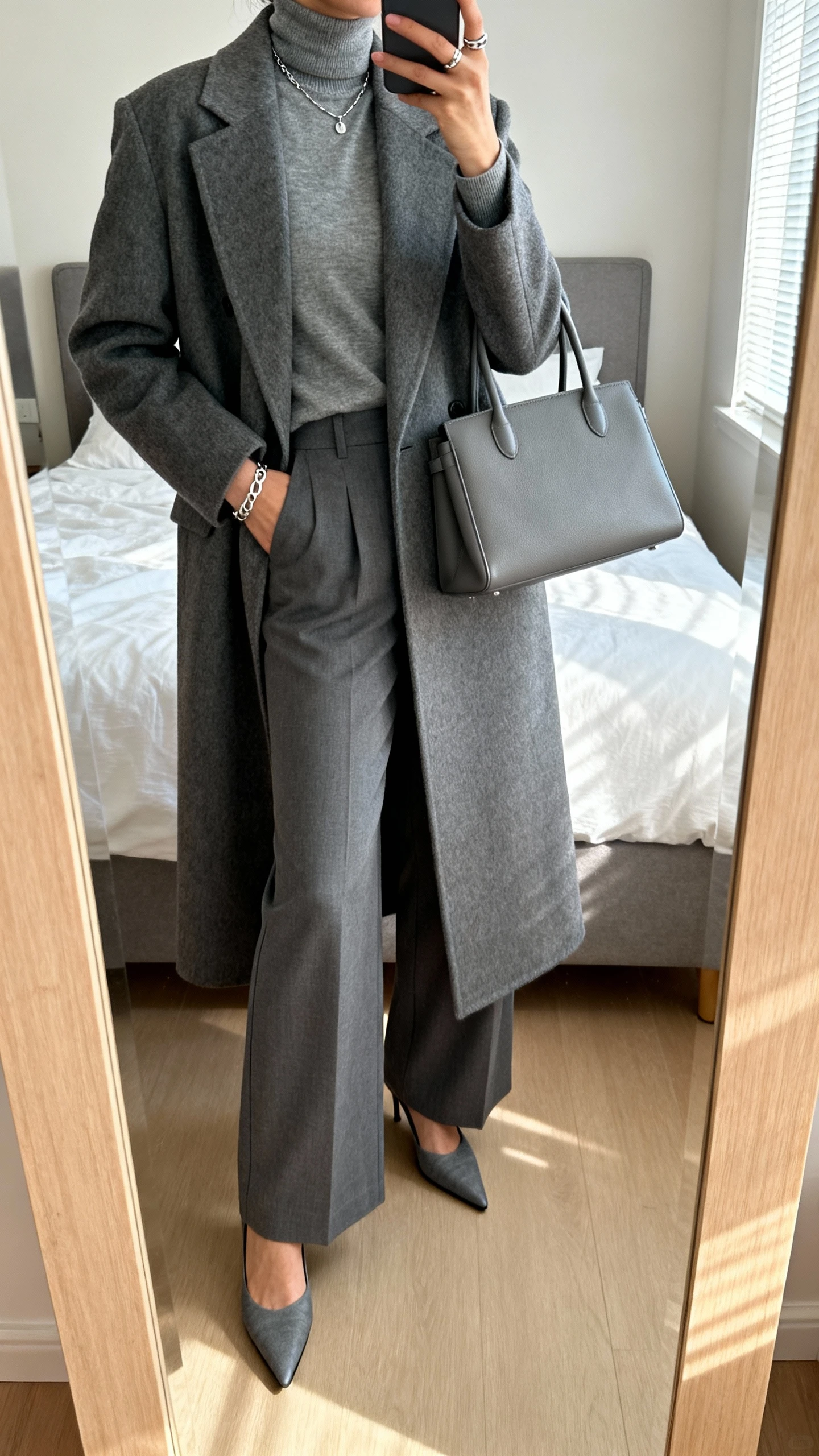 iPhone mirror selfie of a woman wearing a tailored long grey coat over a grey turtleneck, grey wide-leg trousers, pointed grey heels, minimalist silver jewelry, and a structured grey handbag, face not visible, cropped at neck, bedroom mirror, soft morning window light, iPhone photo quality