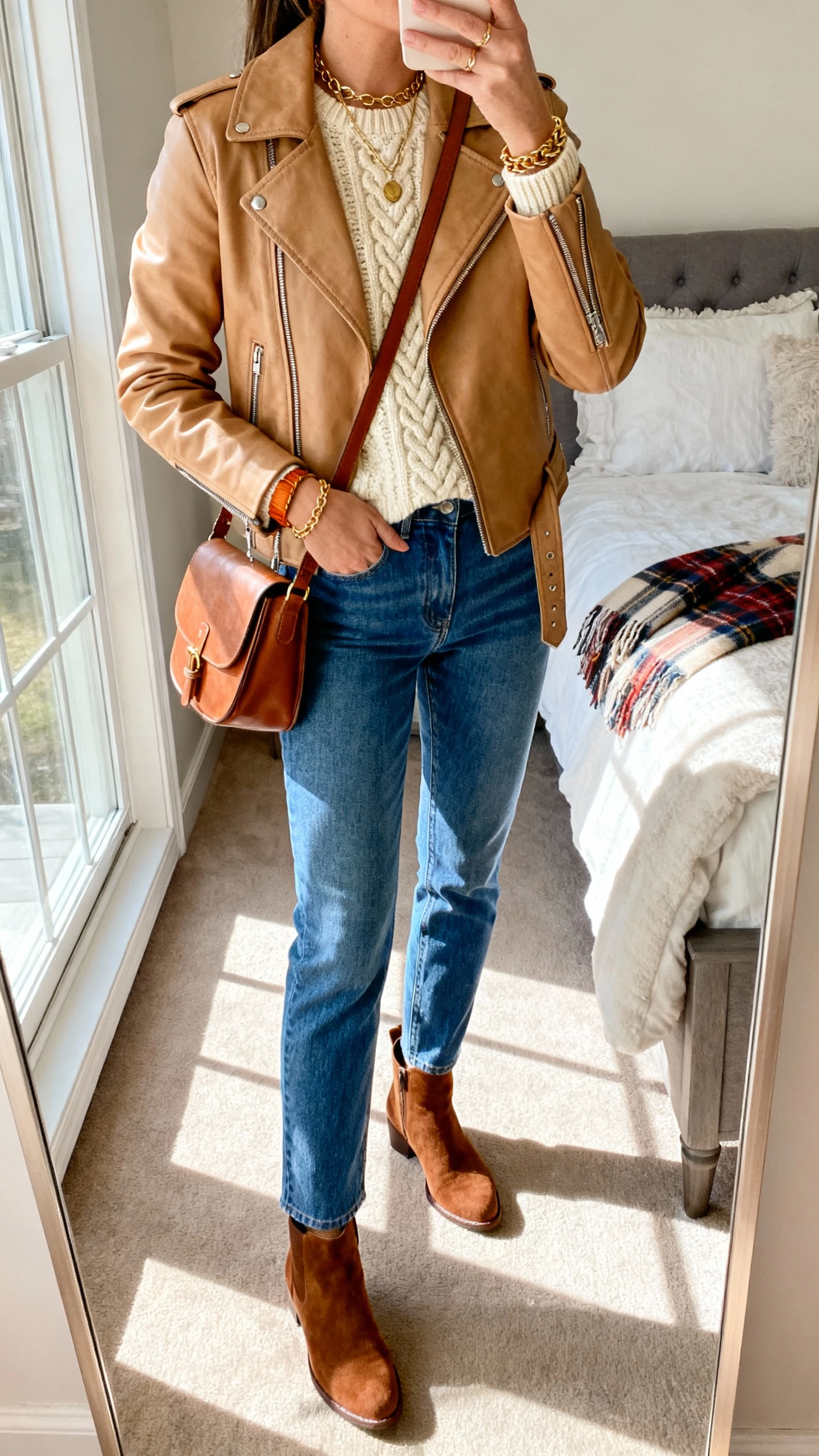 iPhone mirror selfie of a woman wearing a tan leather moto, cream cable-knit sweater, blue straight jeans, brown suede Chelsea boots, cognac crossbody, warm gold jewelry, optional plaid scarf; face not visible, cropped at shoulders, cozy bedroom mirror with afternoon window light, iPhone photo quality.