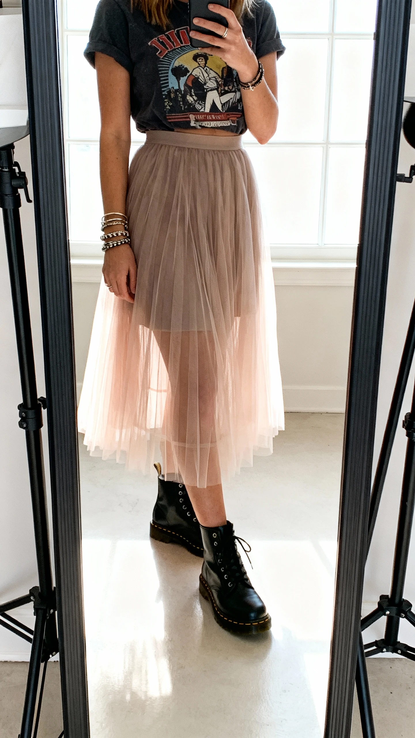 iPhone mirror selfie of a woman wearing a tulle skirt with a tucked-in graphic tee, black Doc Martens, layered bracelets, face not visible, cropped at shoulders, studio-style full-length mirror, bright window light, iPhone photo quality.