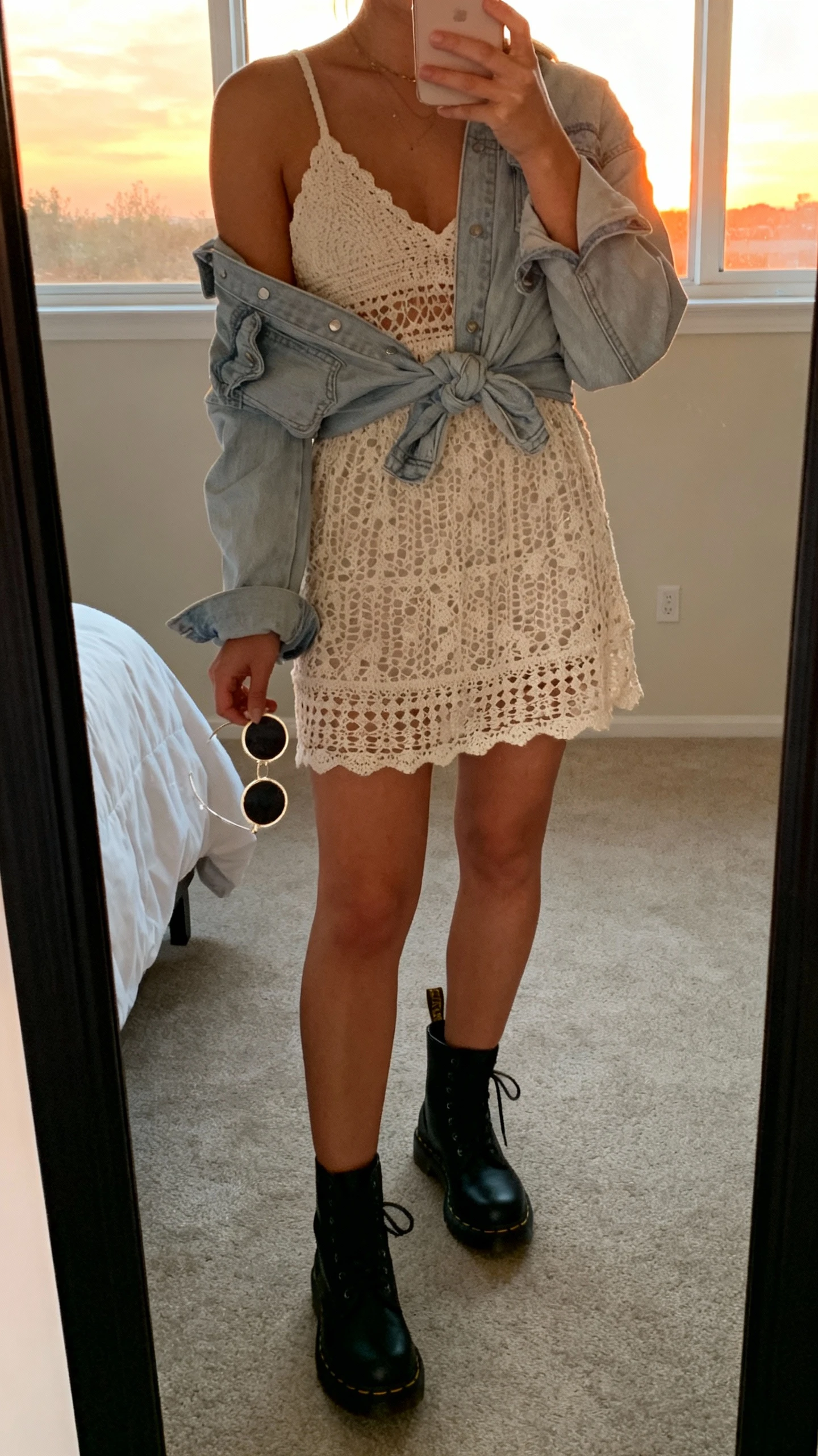 iPhone mirror selfie of a woman wearing an airy crochet dress with black Doc Martens, layered with a light denim jacket tied at the waist and round sunglasses in hand, face not visible, cropped at neck, bedroom mirror, warm sunset window light, iPhone photo quality.