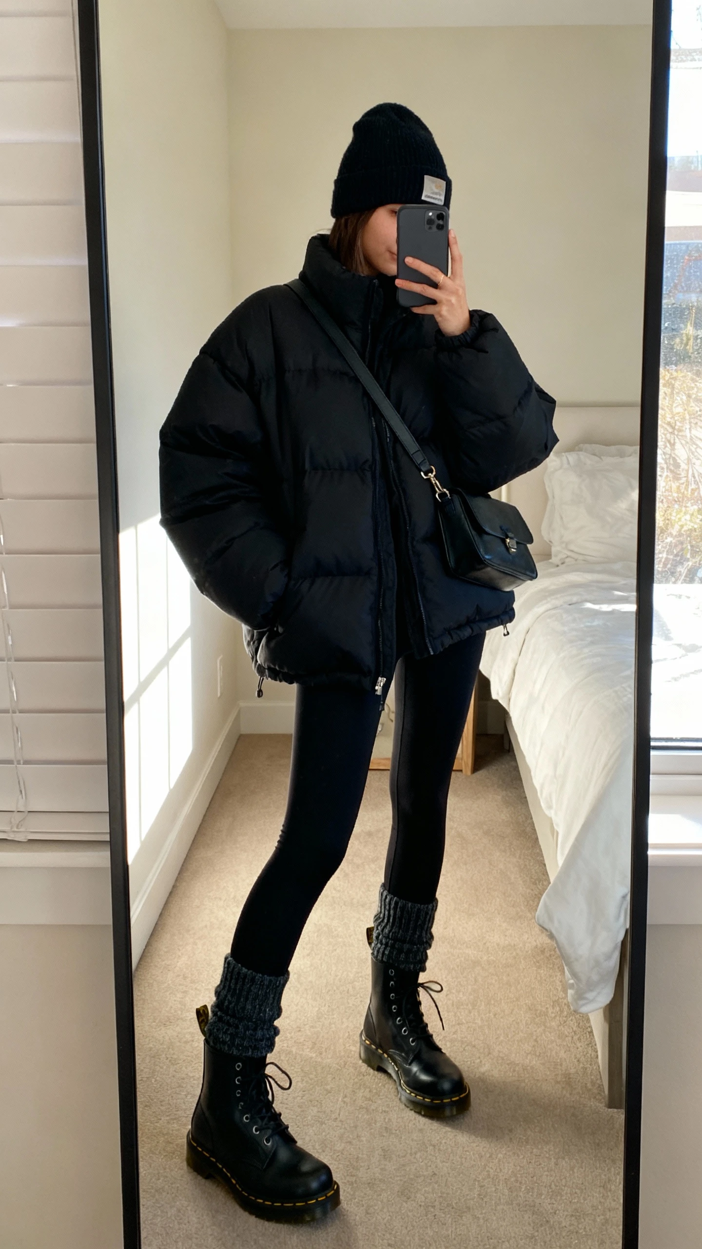 iPhone mirror selfie of a woman wearing an oversized black puffer jacket, black leggings, thick crew socks, black Doc Martens combat boots, a beanie, and a crossbody bag, face not visible, cropped at neck, bedroom mirror with morning window light, iPhone photo quality.