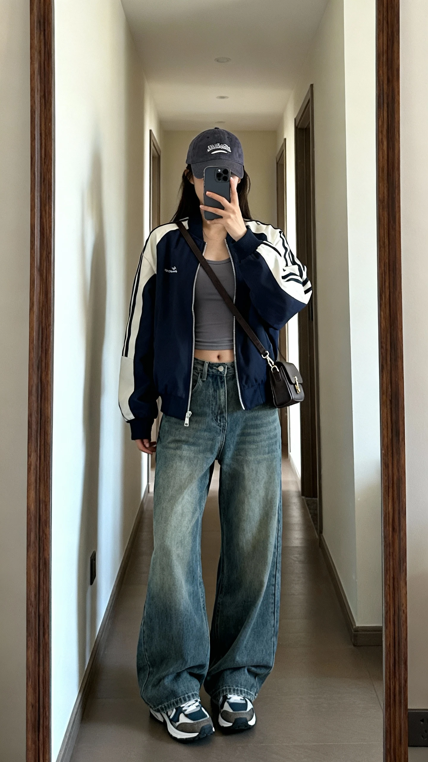iPhone mirror selfie of a woman wearing baggy jeans, a sporty bomber jacket over a fitted tank, baseball cap, retro sneakers, small crossbody bag, face not visible, cropped at shoulders, hallway full-length mirror, natural daylight, iPhone photo quality.