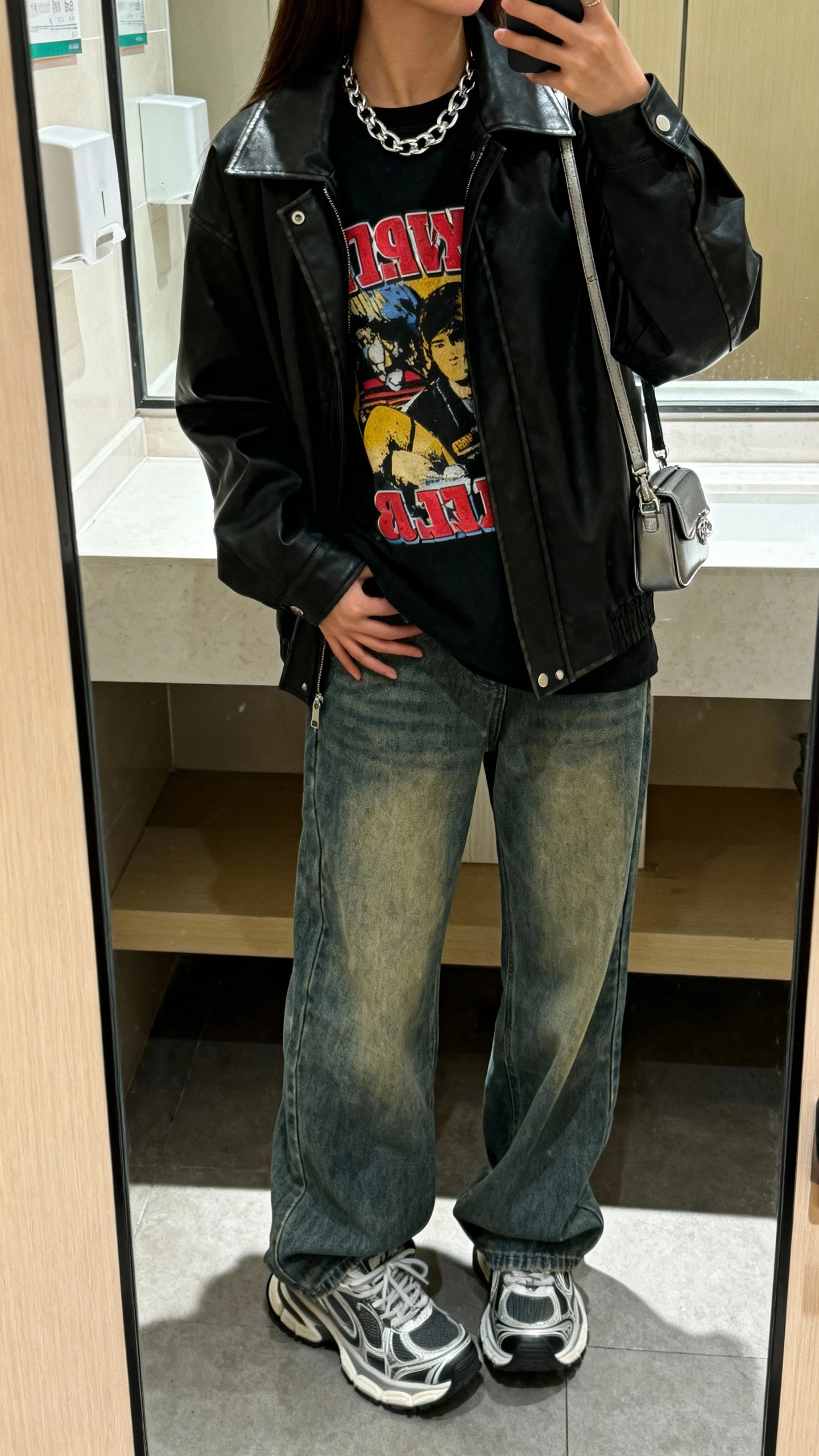 iPhone mirror selfie of a woman wearing baggy jeans, a bold graphic tee under a black leather jacket, chunky sneakers, silver chain necklace, mini shoulder bag, face not visible, cropped at neck, bathroom mirror, indoor lighting, iPhone photo quality.