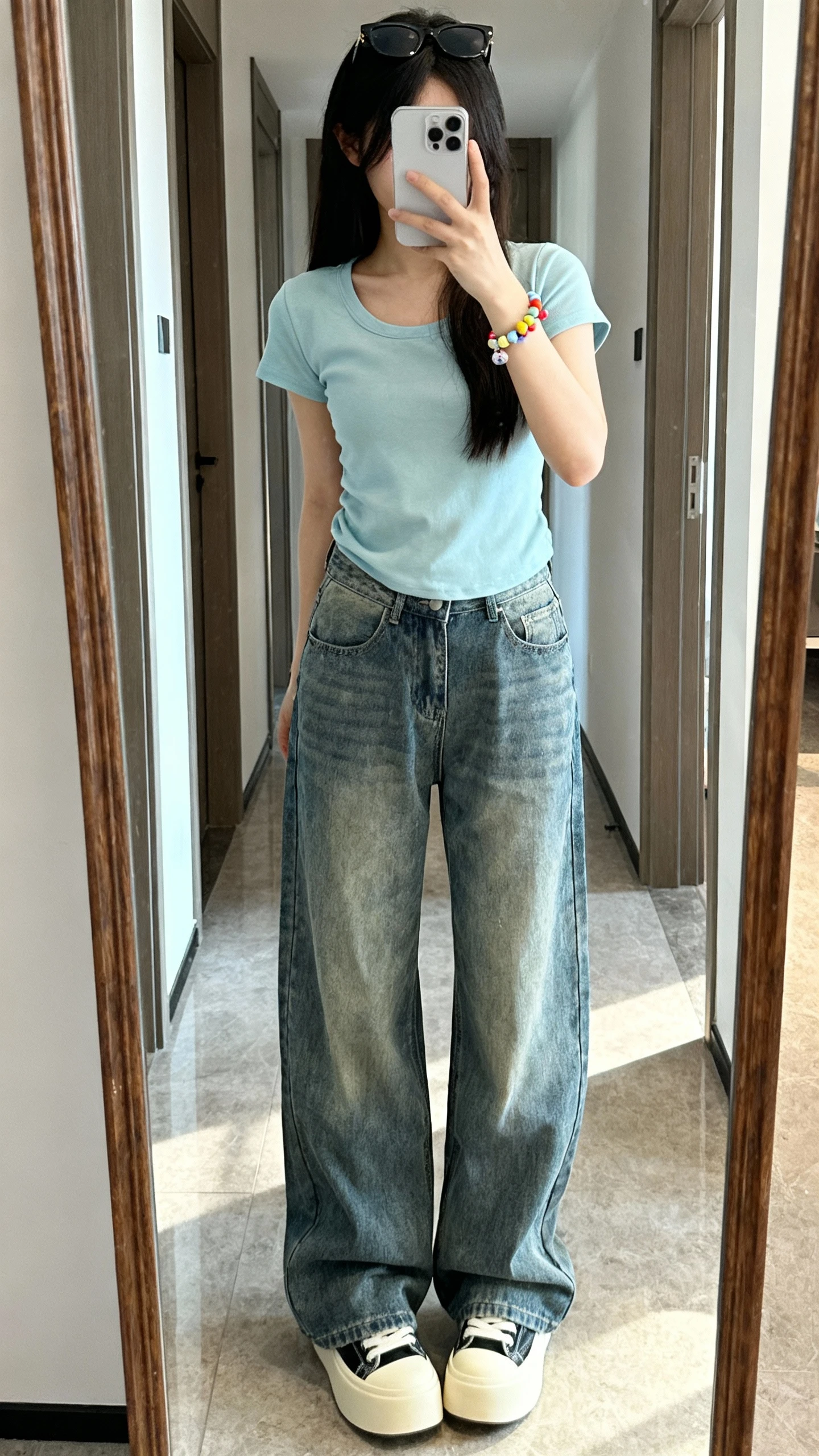 iPhone mirror selfie of a woman wearing baggy jeans, a baby tee, platform sneakers, tiny sunglasses perched on head, playful beaded bracelet, face not visible, cropped at shoulders, hallway mirror, bright daylight, iPhone photo quality.