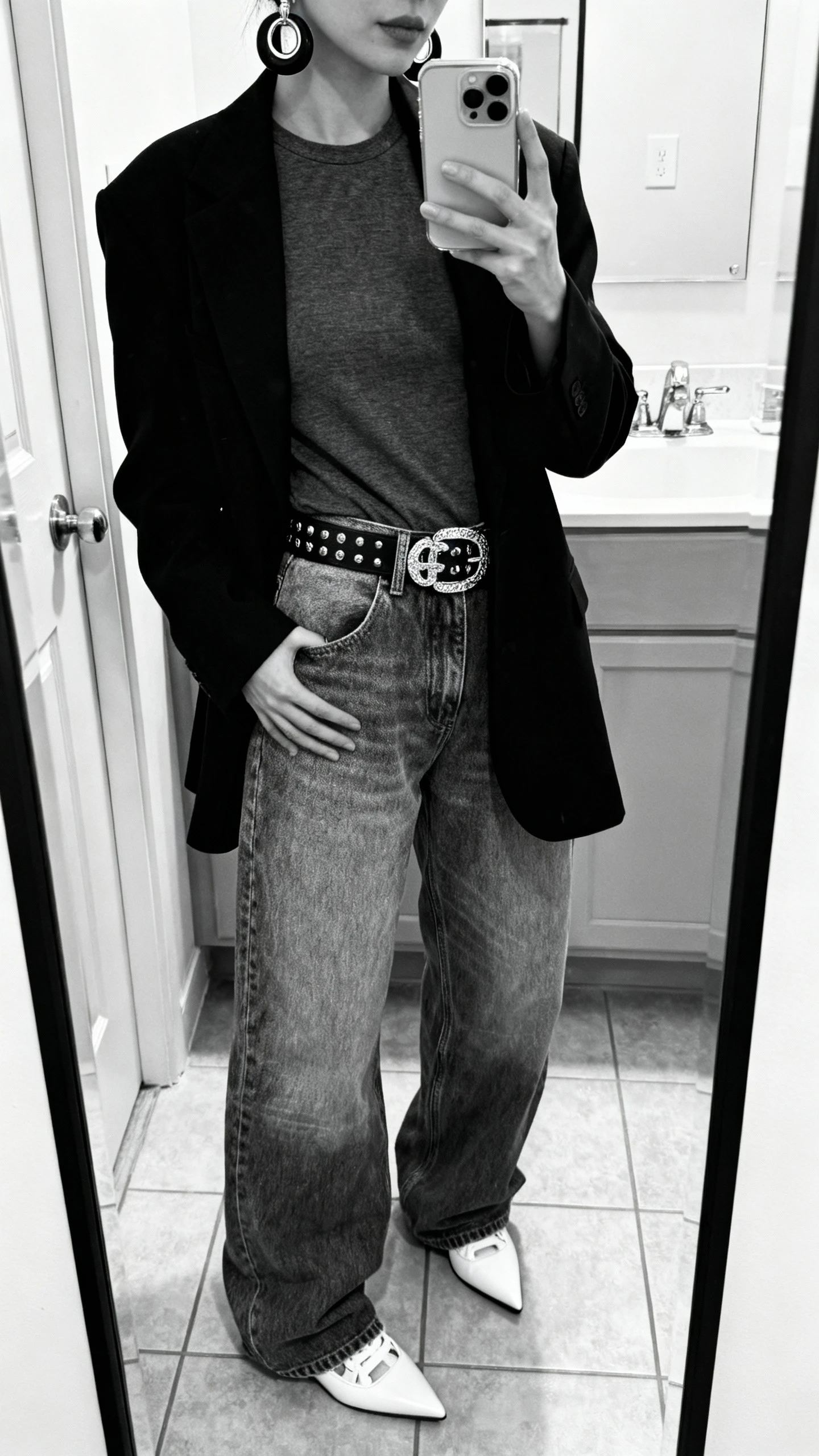 iPhone mirror selfie of a woman wearing baggy jeans in an all-black or all-white monochrome look (matching top and outerwear), statement belt and bold earrings, clean sneakers or pointed flats, face not visible, cropped at neck, bathroom mirror, indoor lighting, iPhone photo quality.