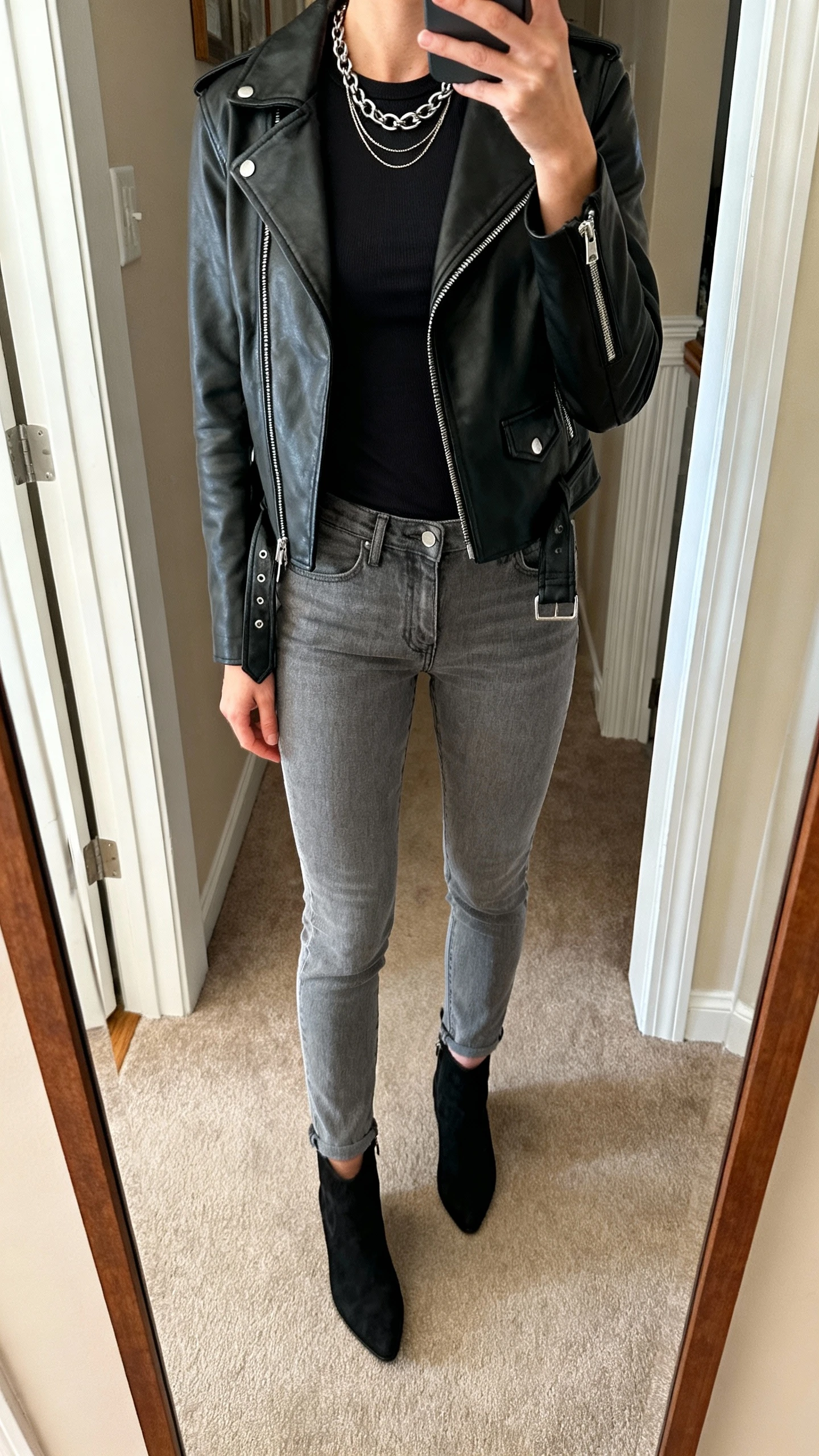 iPhone mirror selfie of a woman wearing grey jeans, a black leather moto jacket over a fitted black tee, black ankle boots, layered chain necklace, face not visible, cropped at neck, entryway mirror, warm indoor lighting, iPhone photo quality.