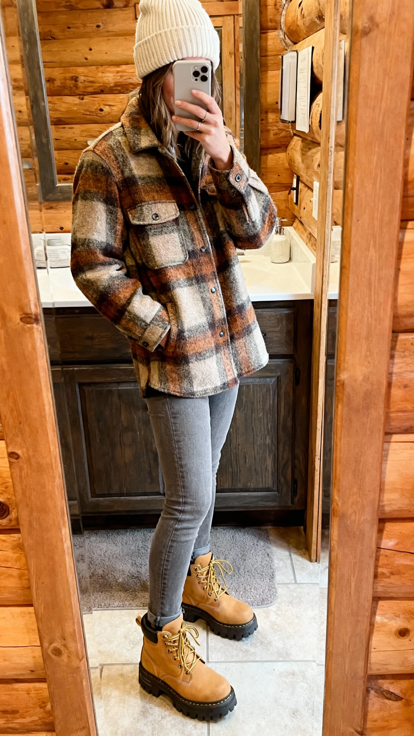 iPhone mirror selfie of a woman wearing grey jeans, a plaid wool shacket in earthy tones, chunky hiking-inspired lace-up boots in tan, ribbed beanie in cream, face not visible, cropped at shoulders, cabin-style bathroom mirror, soft indoor light, iPhone photo quality.