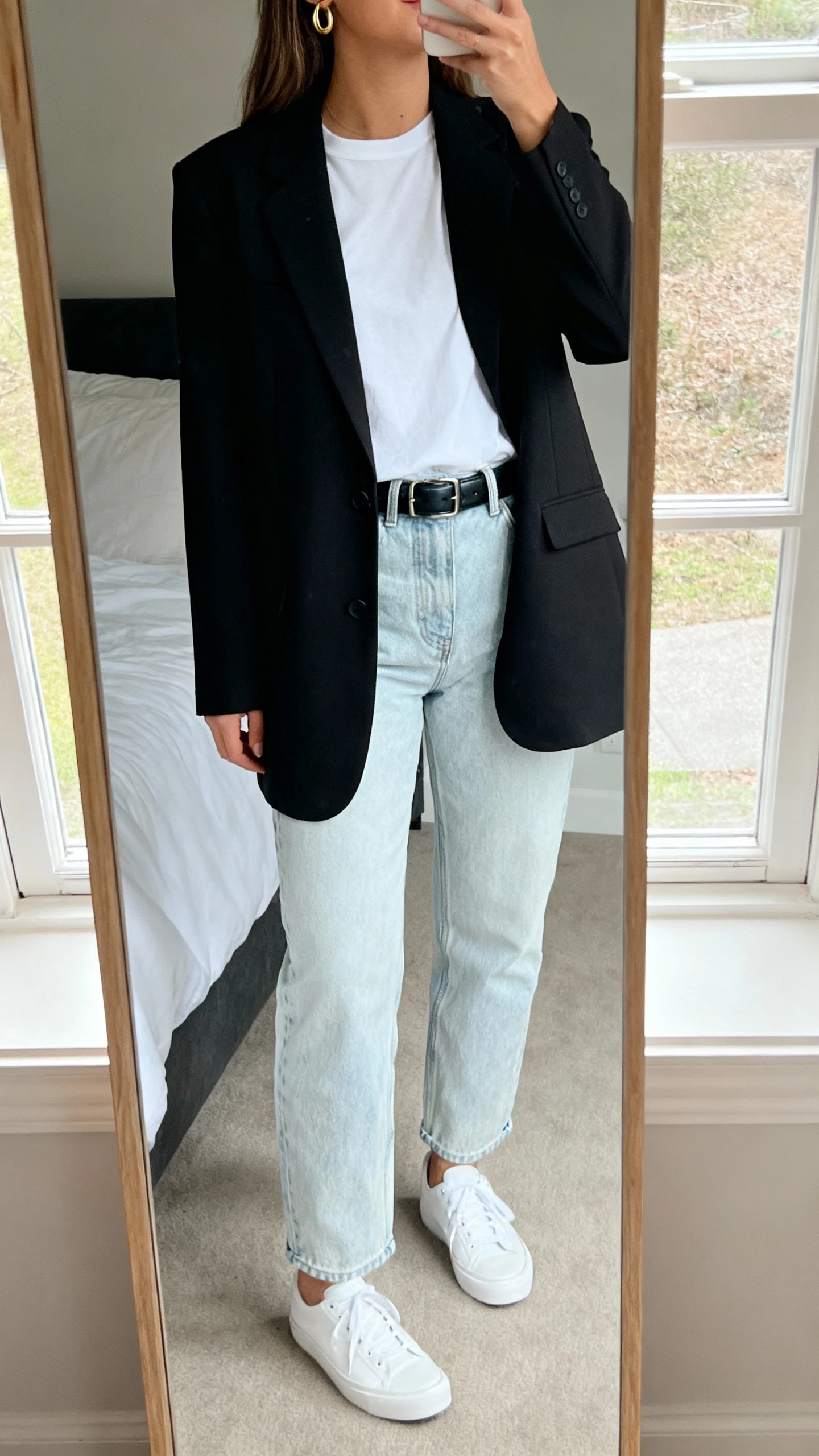 iPhone mirror selfie of a woman wearing light-wash mom jeans, a crisp white tee tucked in, a tailored black blazer, minimal gold hoops, a black leather belt, and white low-top sneakers, face not visible, cropped at neck, bedroom mirror with natural window light, iPhone photo quality.