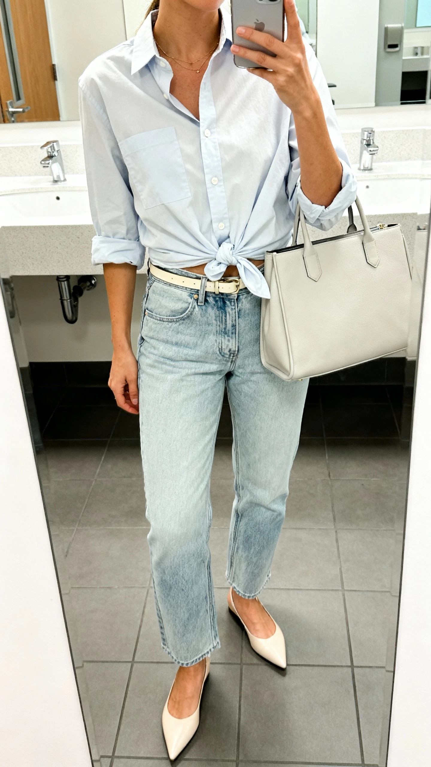 iPhone mirror selfie of a woman wearing light-wash mom jeans, a crisp button-up shirt with a half-tuck and rolled sleeves, pointed-toe flats, a structured tote, and a thin belt, face not visible, cropped at neck, office restroom mirror, bright indoor lighting, iPhone photo quality.