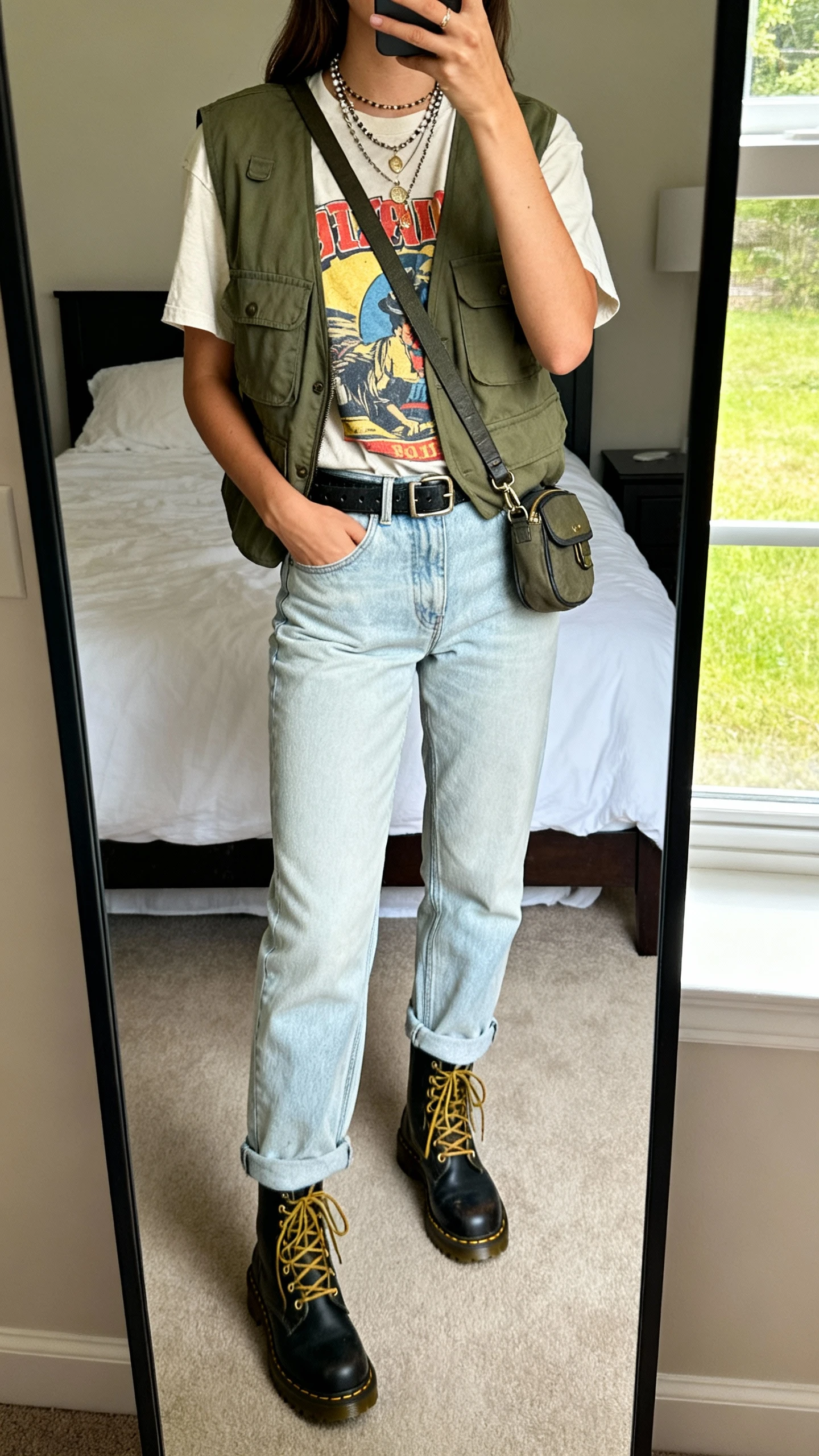 iPhone mirror selfie of a woman wearing light-wash mom jeans, a vintage graphic tee, an olive utility vest, lace-up combat boots, layered necklaces, and a crossbody belt bag, face not visible, full-length mirror in bedroom, afternoon window light, iPhone photo quality.