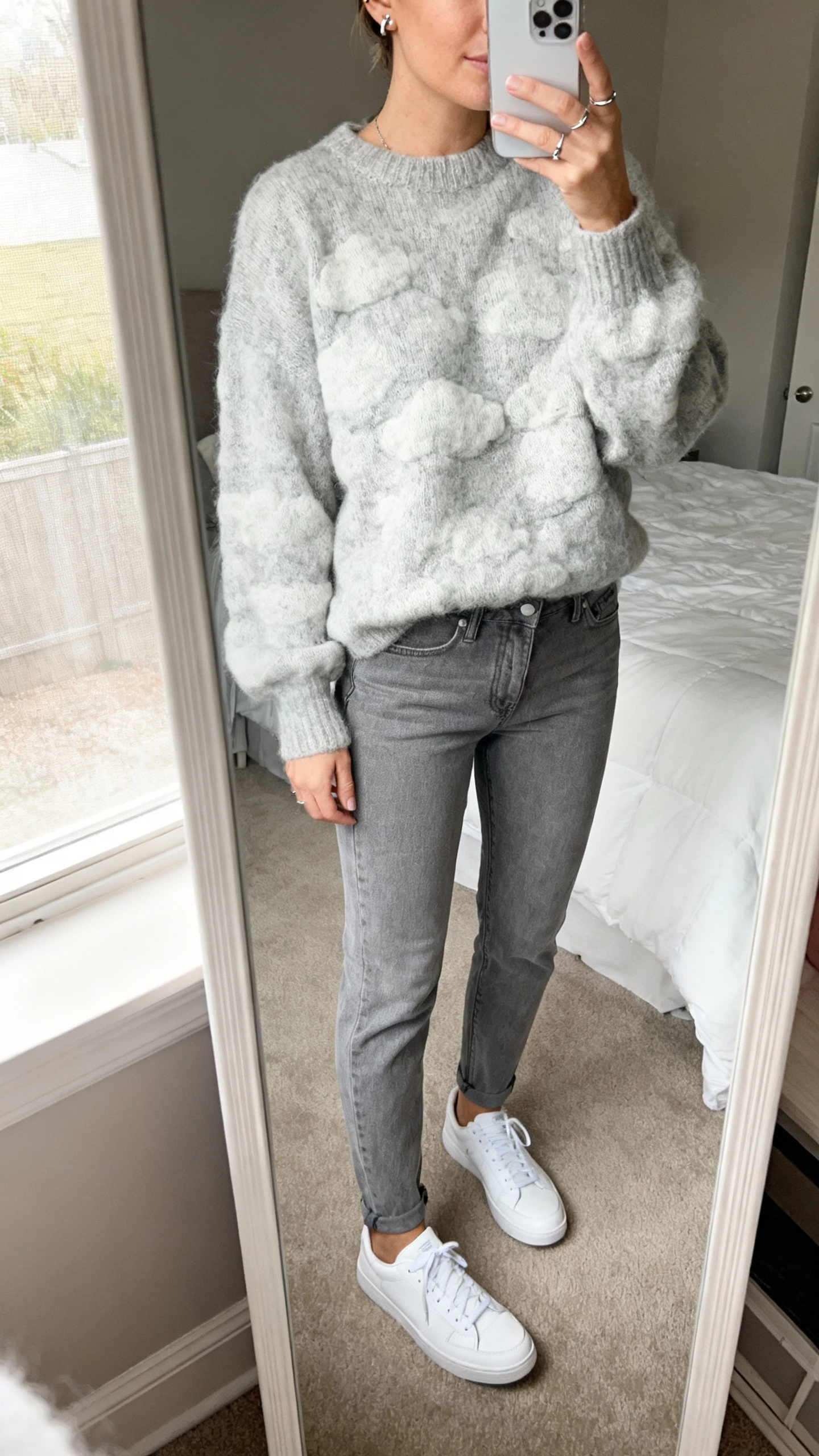 iPhone mirror selfie of a woman wearing soft grey jeans with a cloud-soft light grey knit sweater, clean white low-top sneakers, minimal silver studs, face not visible, cropped at neck, bedroom mirror, natural window light, iPhone photo quality.