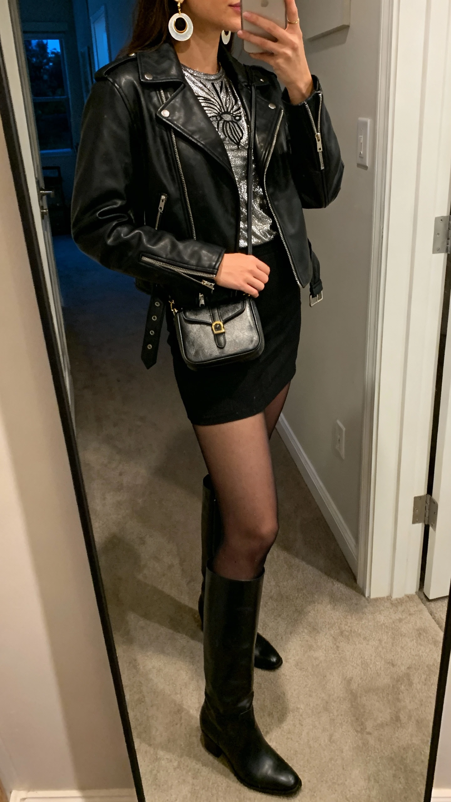 iPhone side-angle mirror selfie of a woman in a classic black leather moto, metallic statement top, black mini skirt, knee-high boots, compact shoulder bag, bold earrings; face not visible, hallway full-length mirror, night indoor lighting, optional sheer black tights, iPhone photo quality.