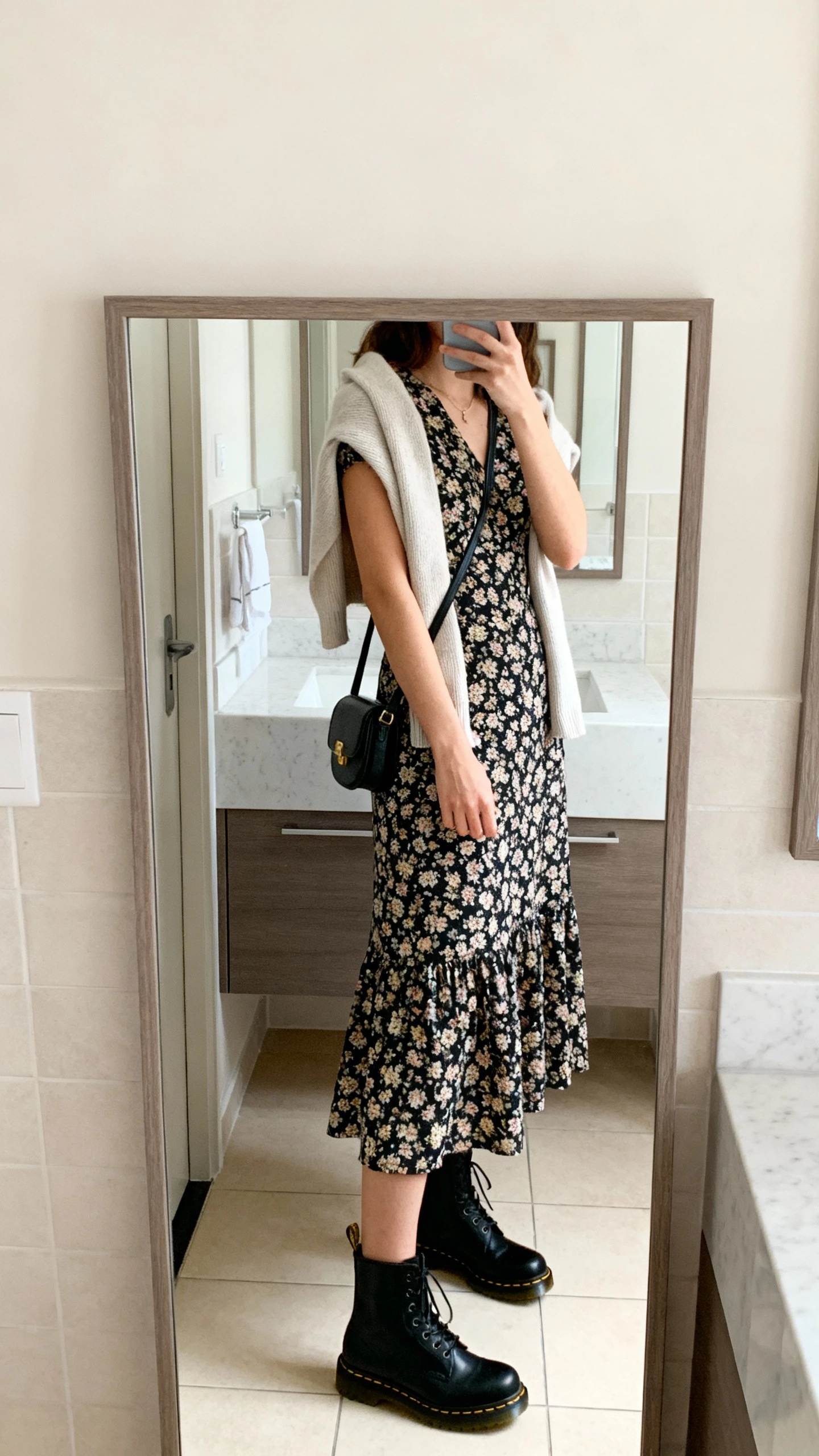 iPhone side-angle mirror selfie of a woman in a floral midi dress with classic black Doc Martens 1460 boots, light cardigan draped over shoulders and a small crossbody bag, face not visible, bathroom full-length mirror, soft morning light, iPhone photo quality.