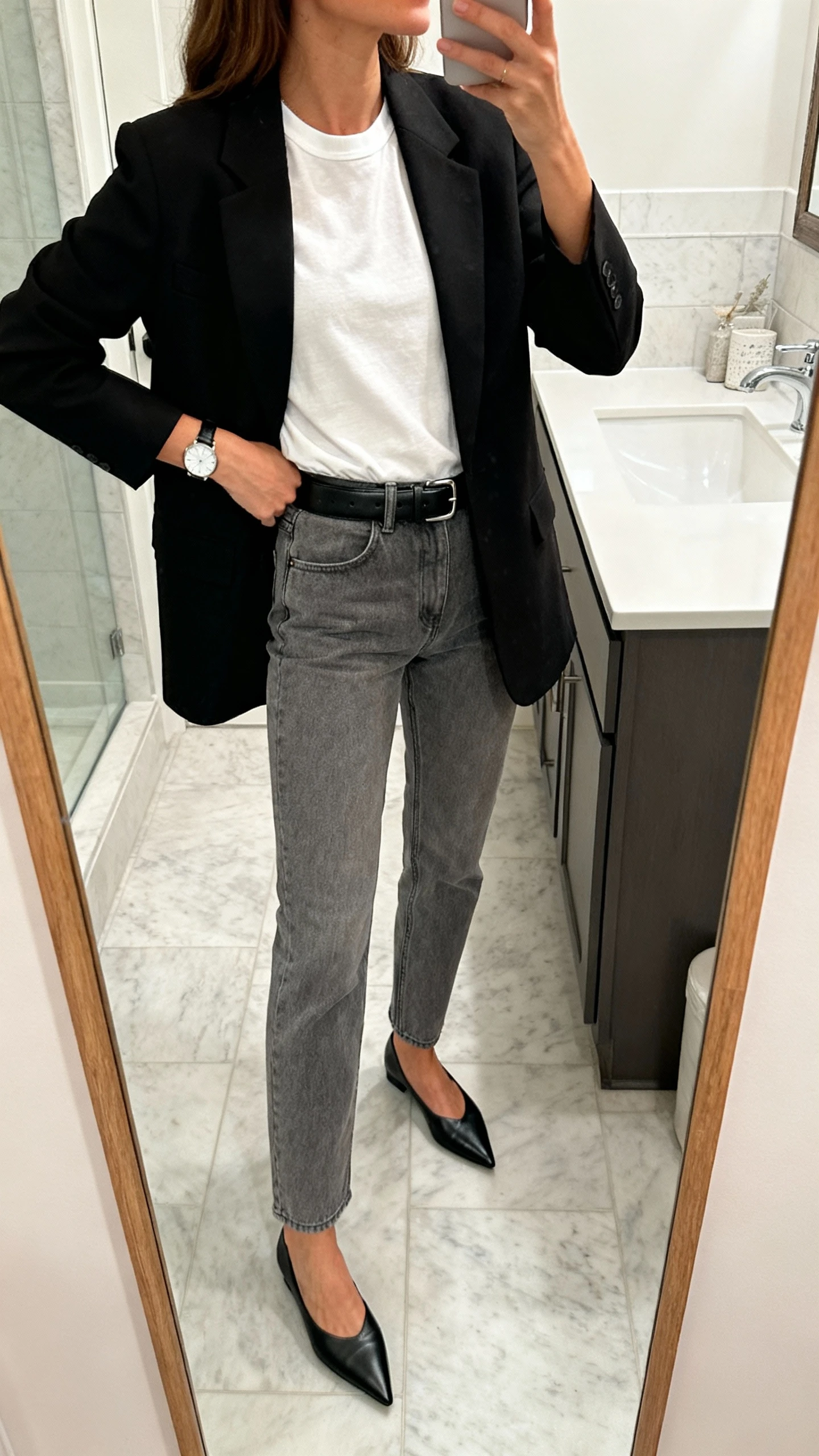 iPhone side-angle mirror selfie of a woman in grey jeans, a sharp black blazer over a white crewneck tee, black leather belt, black pointed flats, simple watch, face not visible, bathroom full-length mirror, morning indoor light, iPhone photo quality.
