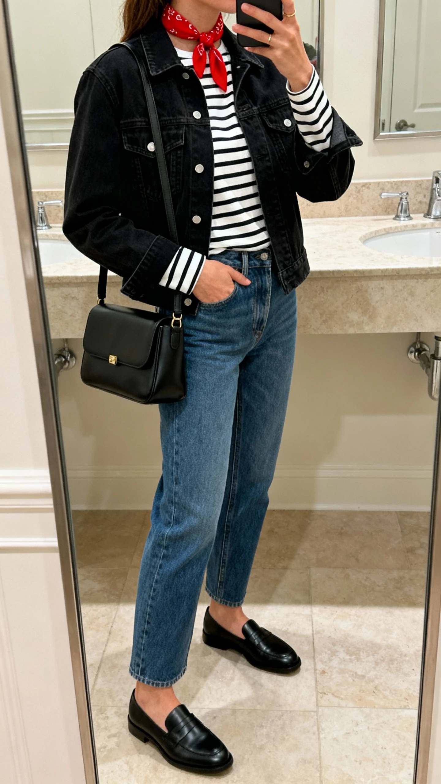iPhone side-angle mirror selfie of a woman wearing a black denim jacket over a black-and-white Breton striped long-sleeve tee, straight-leg blue jeans, black loafers, a red mini neck scarf, and a black structured shoulder bag, face not visible, bathroom mirror, soft indoor lighting, iPhone photo quality.