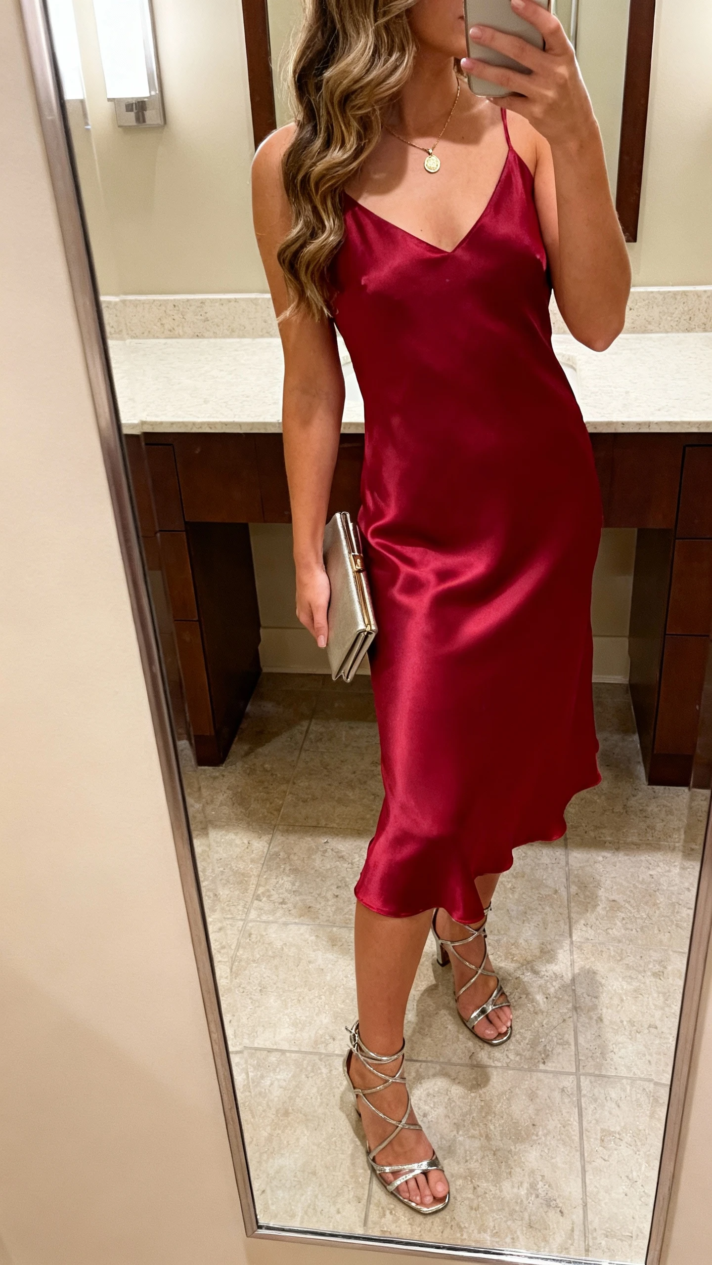 iPhone side-angle mirror selfie of a woman wearing a cherry-red satin slip dress, strappy metallic heels, delicate pendant necklace, slim clutch, soft waves over shoulders, face not visible, bathroom mirror, warm indoor lighting, iPhone photo quality