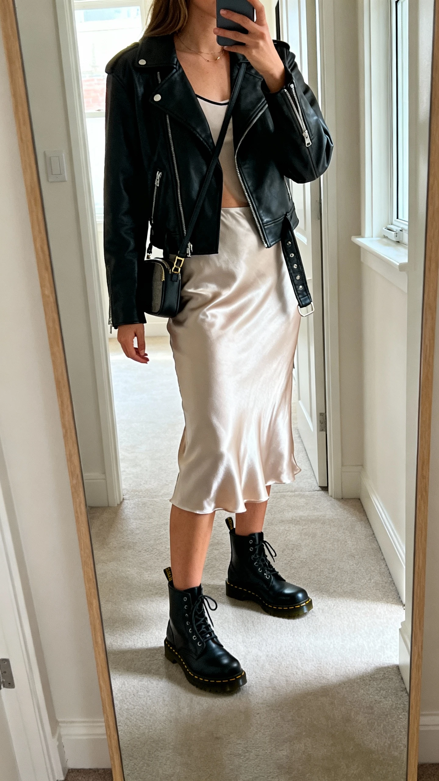 iPhone side-angle mirror selfie of a woman wearing a satin slip skirt, a black leather moto jacket over a simple camisole, black Doc Martens and a small crossbody, face not visible, cropped at shoulders, hallway full-length mirror, natural window light, iPhone photo quality.