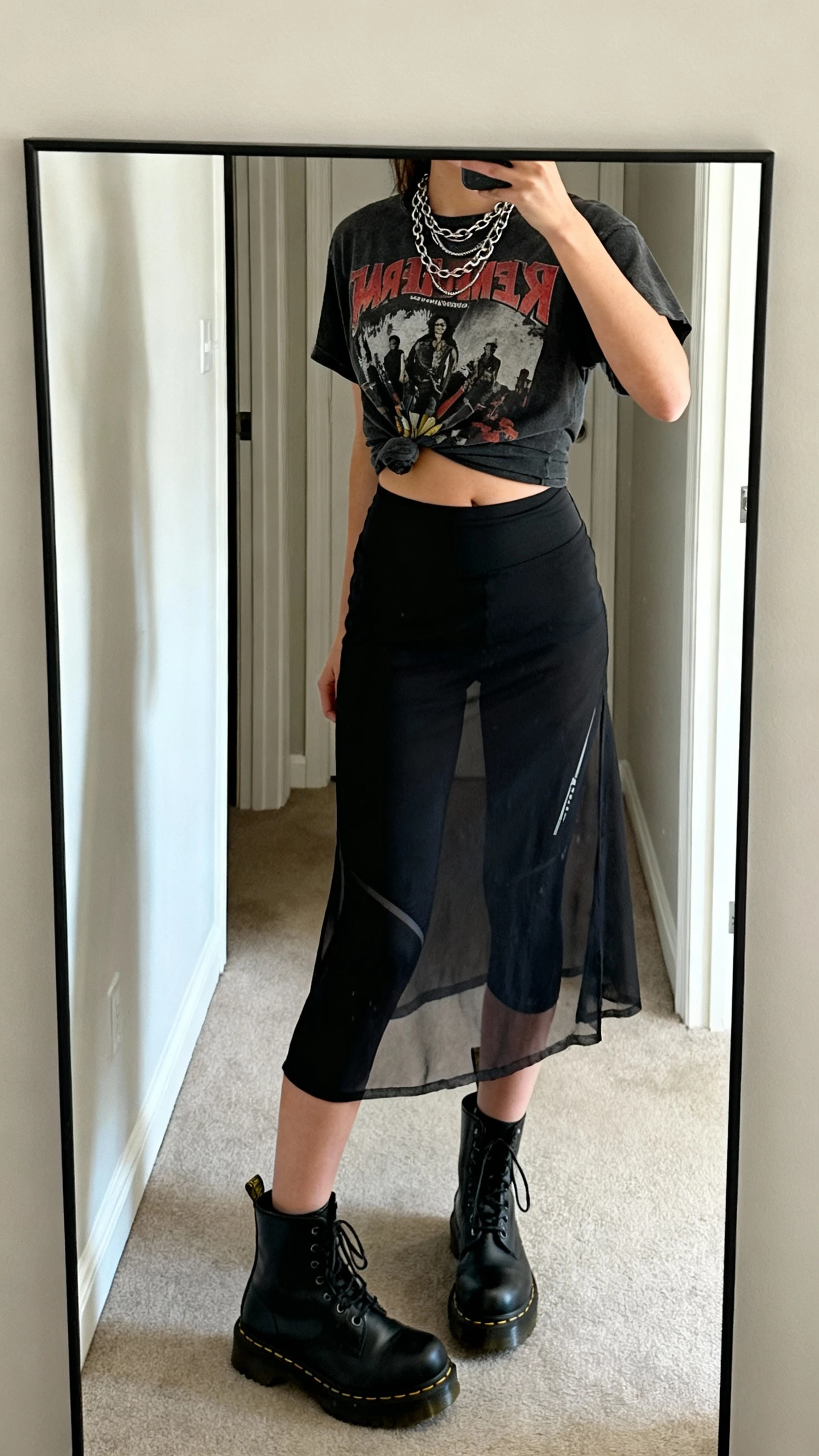 iPhone side-angle mirror selfie of a woman wearing a sheer black midi skirt over biker shorts, vintage band tee half-tucked, chunky black Doc Martens, layered chain necklaces, face not visible, full-length mirror in hallway, natural daylight, iPhone photo quality.