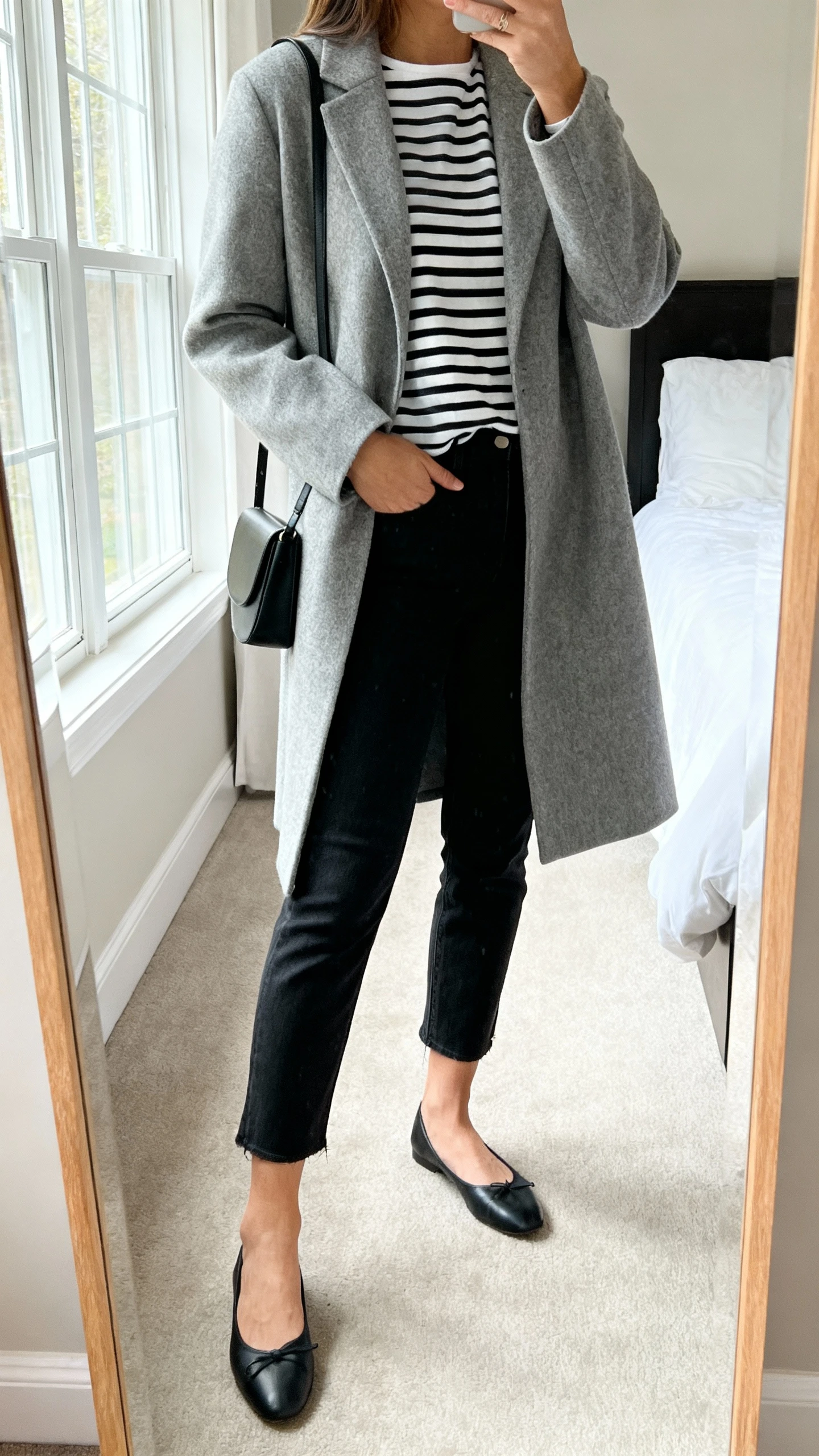 iPhone side-angle mirror selfie of a woman wearing a grey coat over a black-and-white Breton striped top, black ankle-grazing jeans, black ballet flats, and a slim black shoulder bag, face not visible, cropped at shoulders, bedroom mirror, natural window light, iPhone photo quality