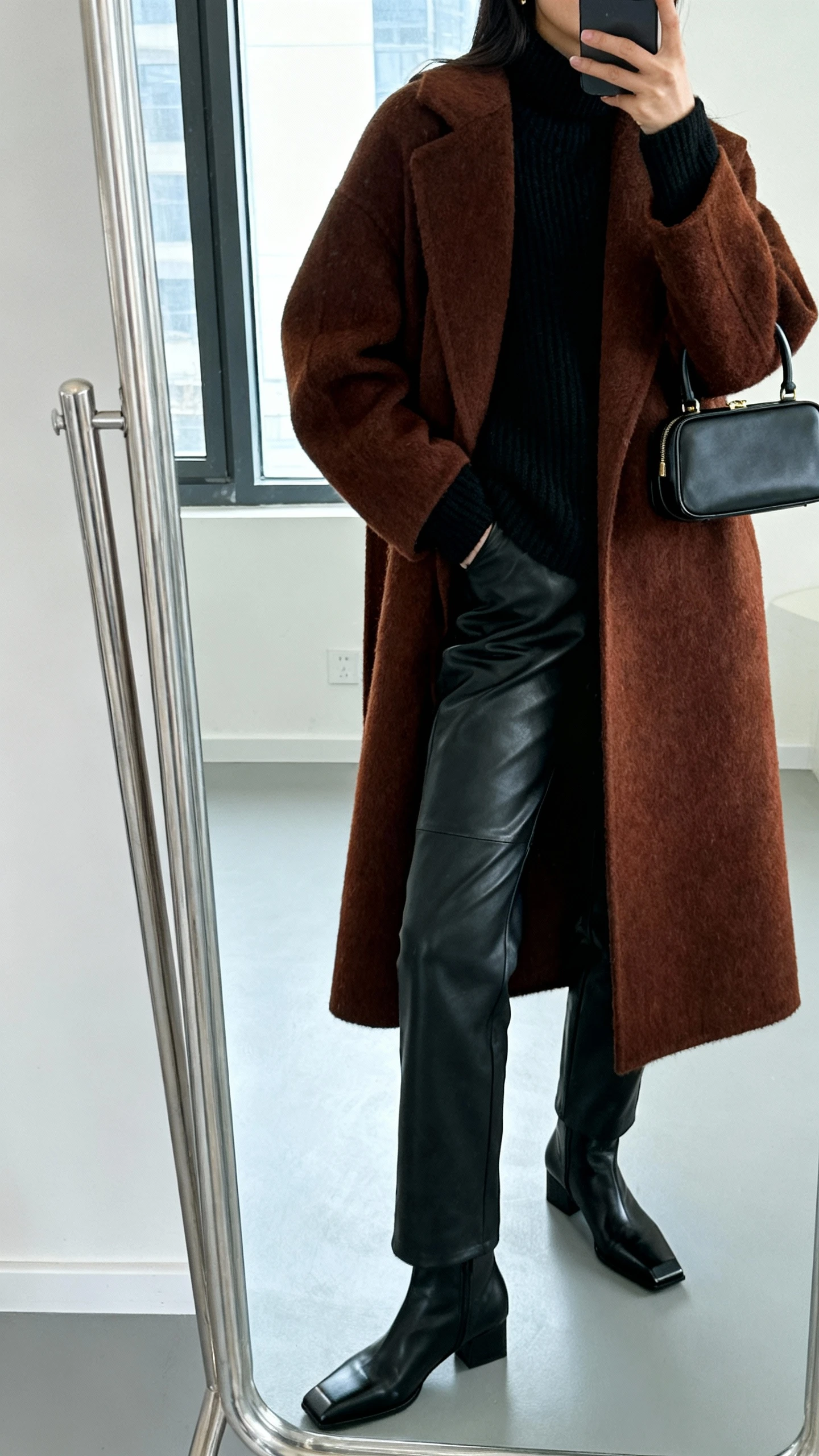 iPhone side-angle mirror selfie of a woman wearing a rich brown wool coat with black leather pants, black cashmere sweater, black square-toe ankle boots, and a mini top-handle bag, face not visible, cropped at neck, full-length mirror, cool window light, iPhone photo quality.