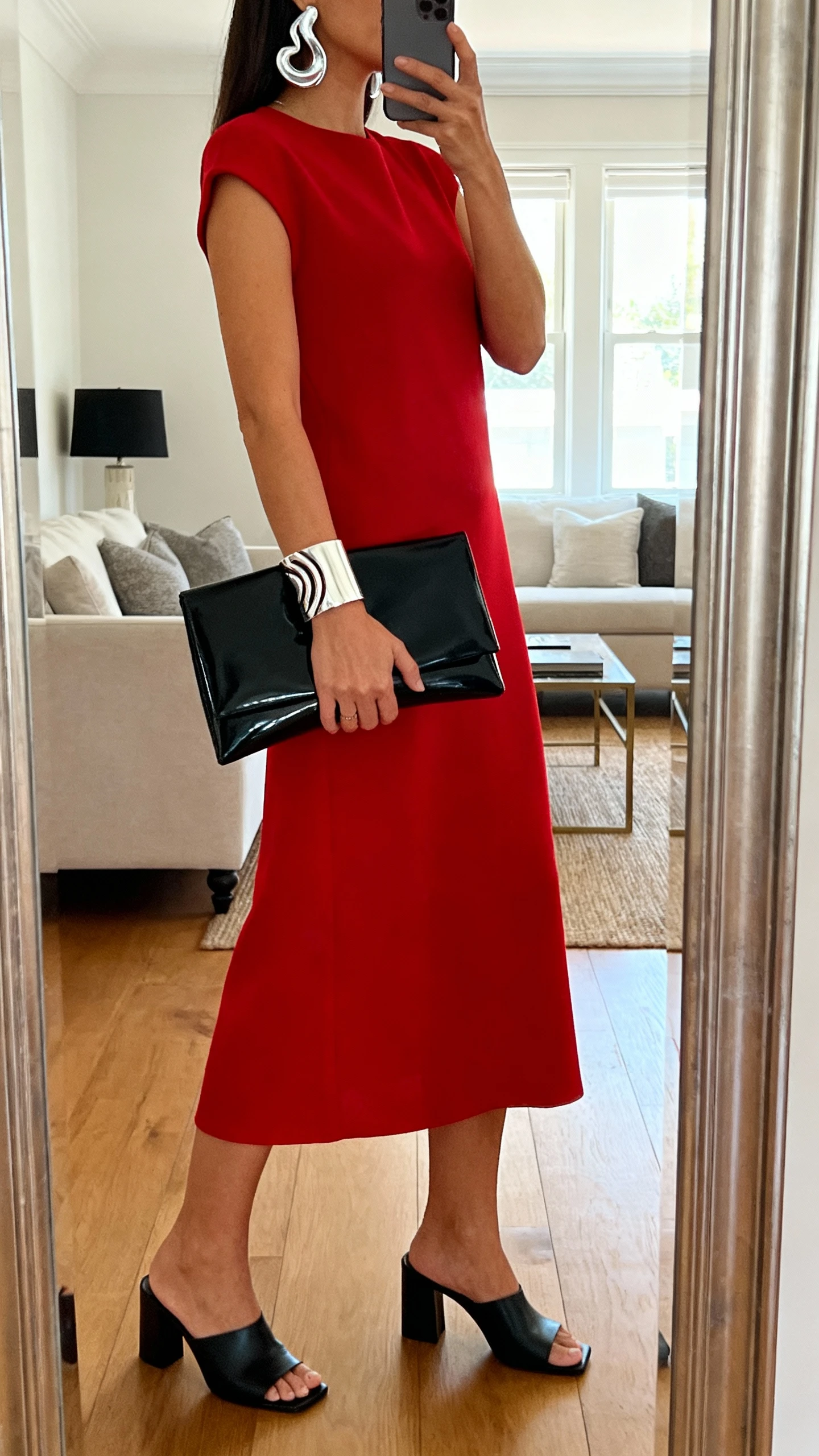 iPhone side-angle mirror selfie of a woman wearing a minimalist red column dress, architectural silver cuff bracelet, sculptural silver earrings, sleek black clutch, black heeled mules, face not visible, living room mirror, indirect window light, iPhone photo quality