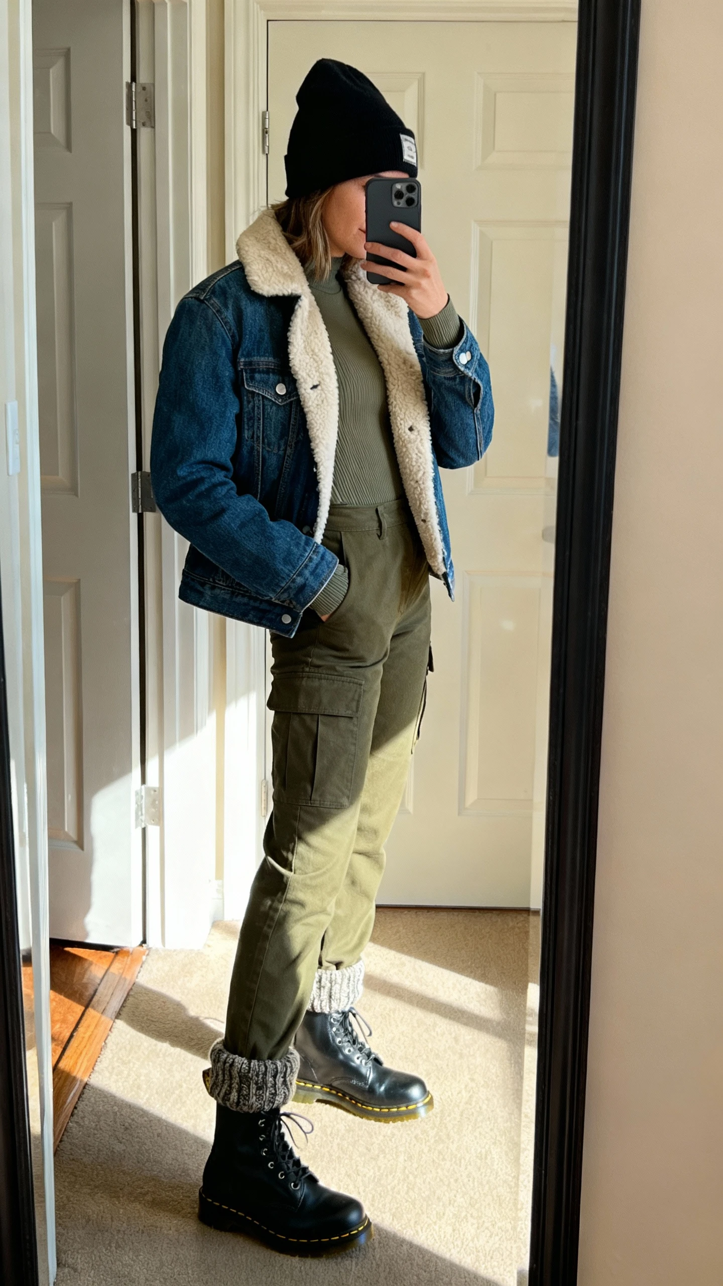 iPhone side-angle mirror selfie of a woman wearing olive utility cargo pants, a sherpa-lined denim jacket over a thermal long-sleeve, beanie, black Doc Martens boots, and chunky wool socks peeking out, face not visible, entryway mirror with afternoon light, iPhone photo quality.