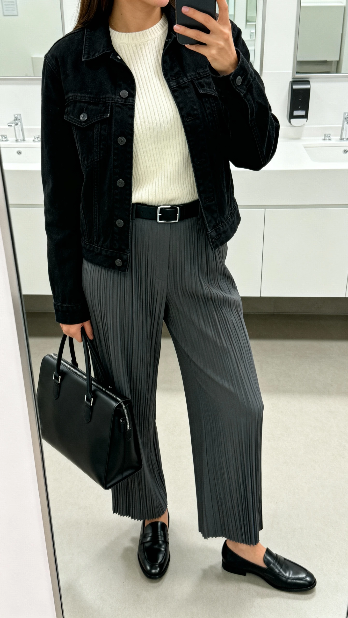 iPhone side-angle mirror selfie of a woman wearing a black denim jacket with charcoal pleated trousers, a cream ribbed crewneck, black leather loafers, a slim black belt, and a structured black work tote, face not visible, office bathroom mirror, bright indoor lighting, iPhone photo quality.