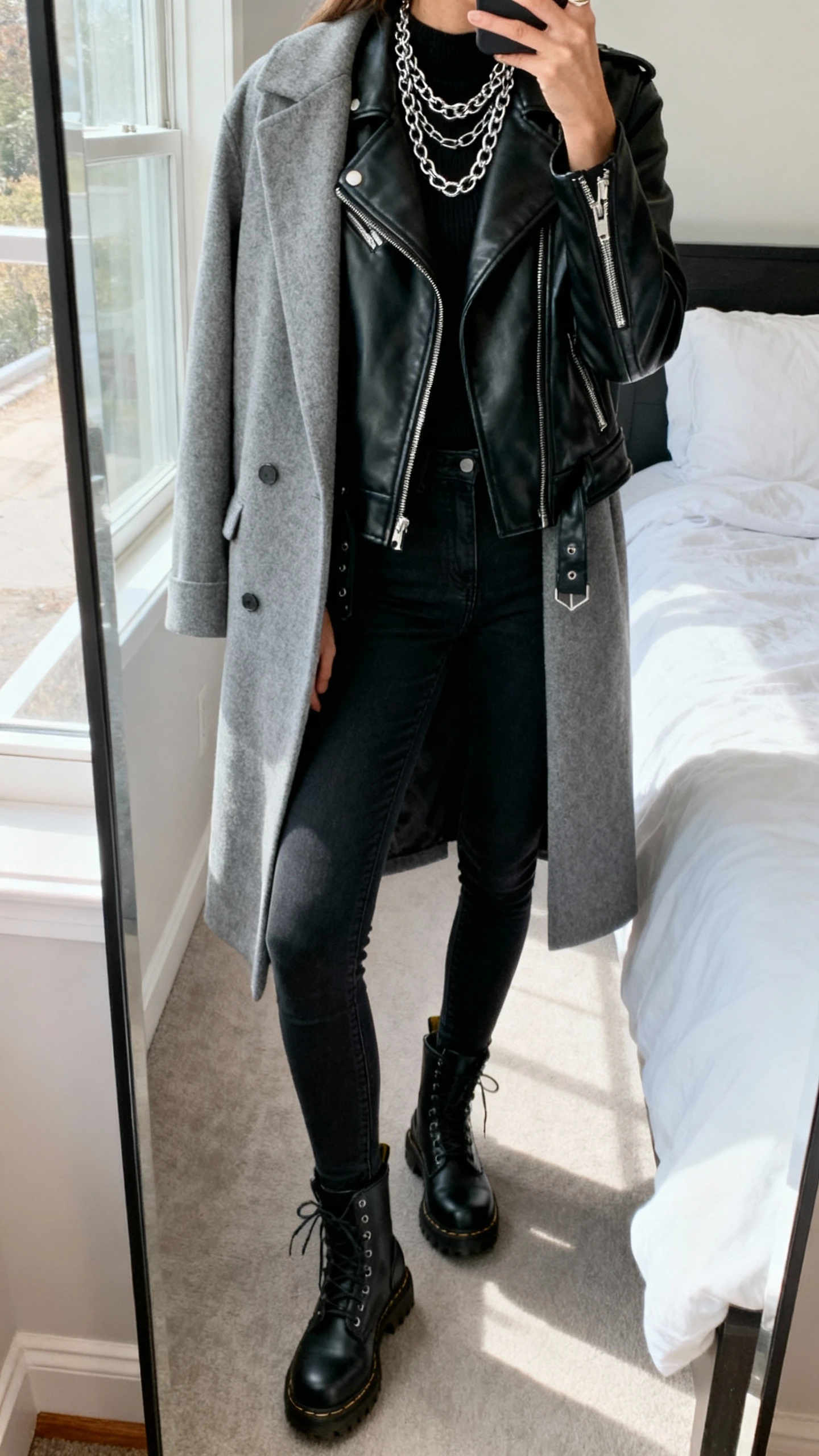 iPhone side-angle mirror selfie of a woman wearing a grey coat layered over a black leather moto jacket, skinny black jeans, black combat boots, and layered silver chains, face not visible, cropped at shoulders, bedroom mirror, natural window light, iPhone photo quality
