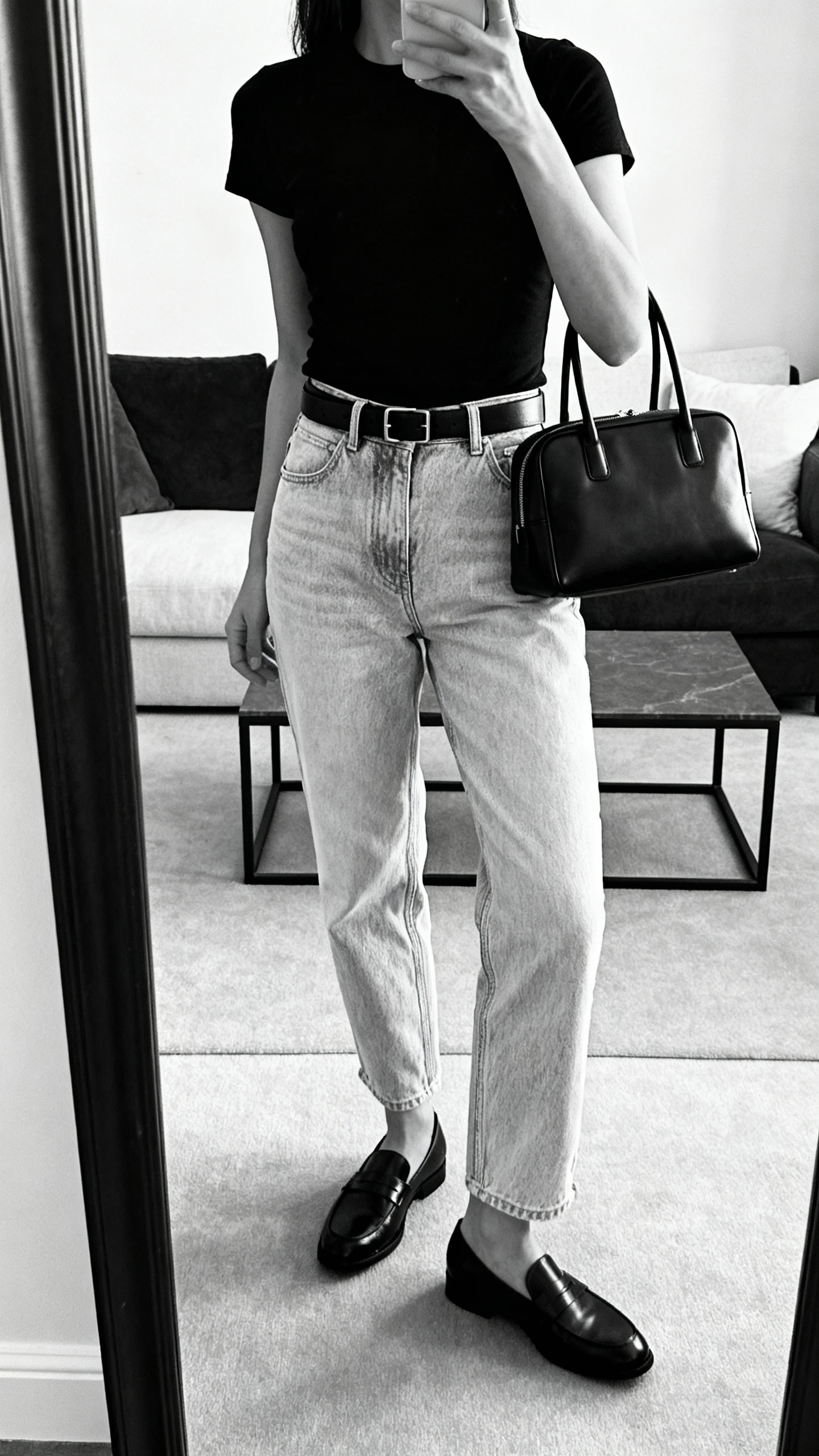 iPhone side-angle mirror selfie of a woman wearing light-wash mom jeans with a black fitted tee, black minimalist belt, black sleek loafers, and a black structured handbag for a monochrome look, face not visible, cropped at shoulders, living room mirror with soft natural light, iPhone photo quality.
