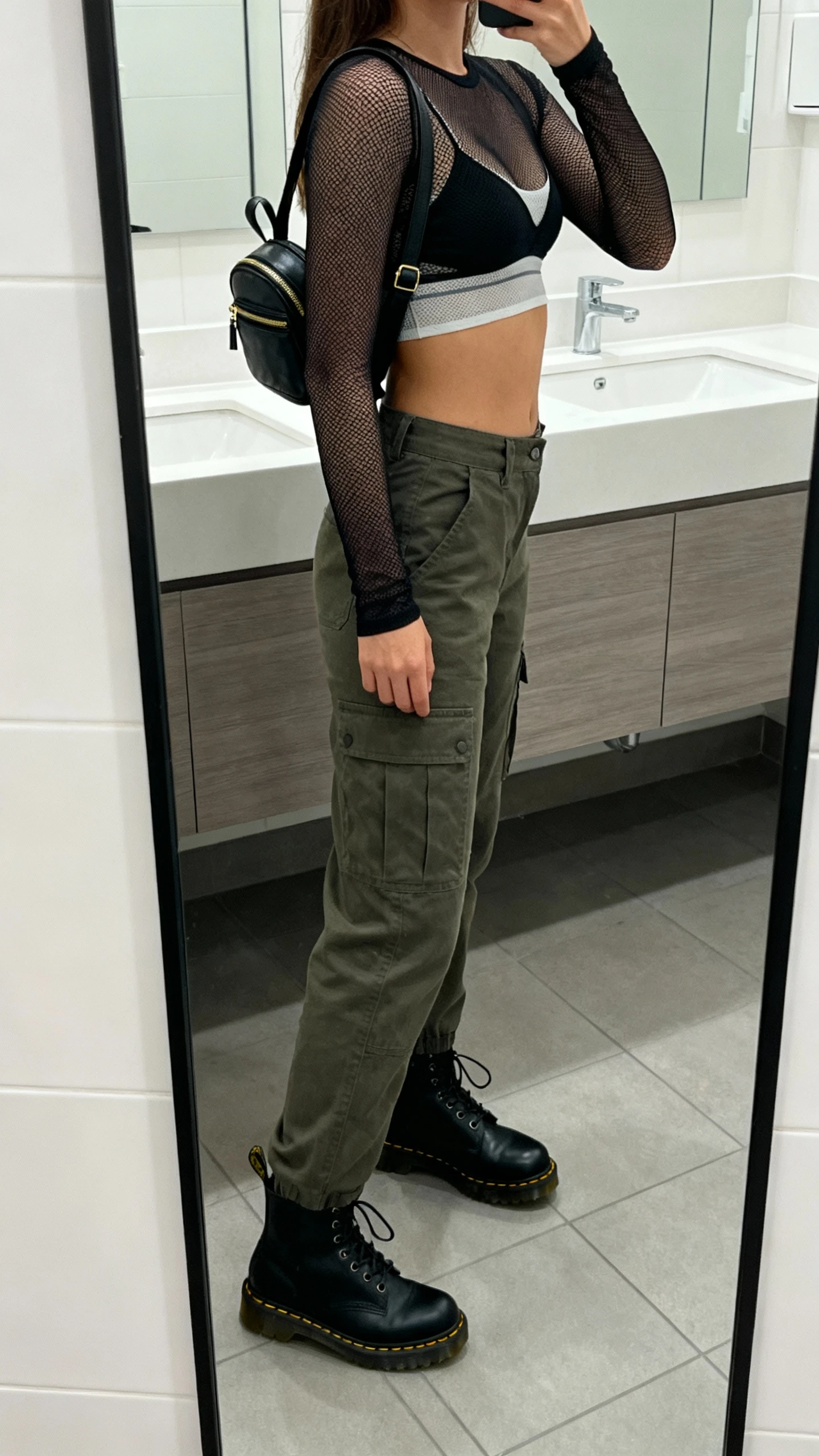 iPhone side-angle mirror selfie of a woman wearing cargo pants with a fitted long-sleeve mesh top over a simple bralette, black Doc Martens, and a mini backpack, face not visible, bathroom mirror, cool indoor lighting, iPhone photo quality.