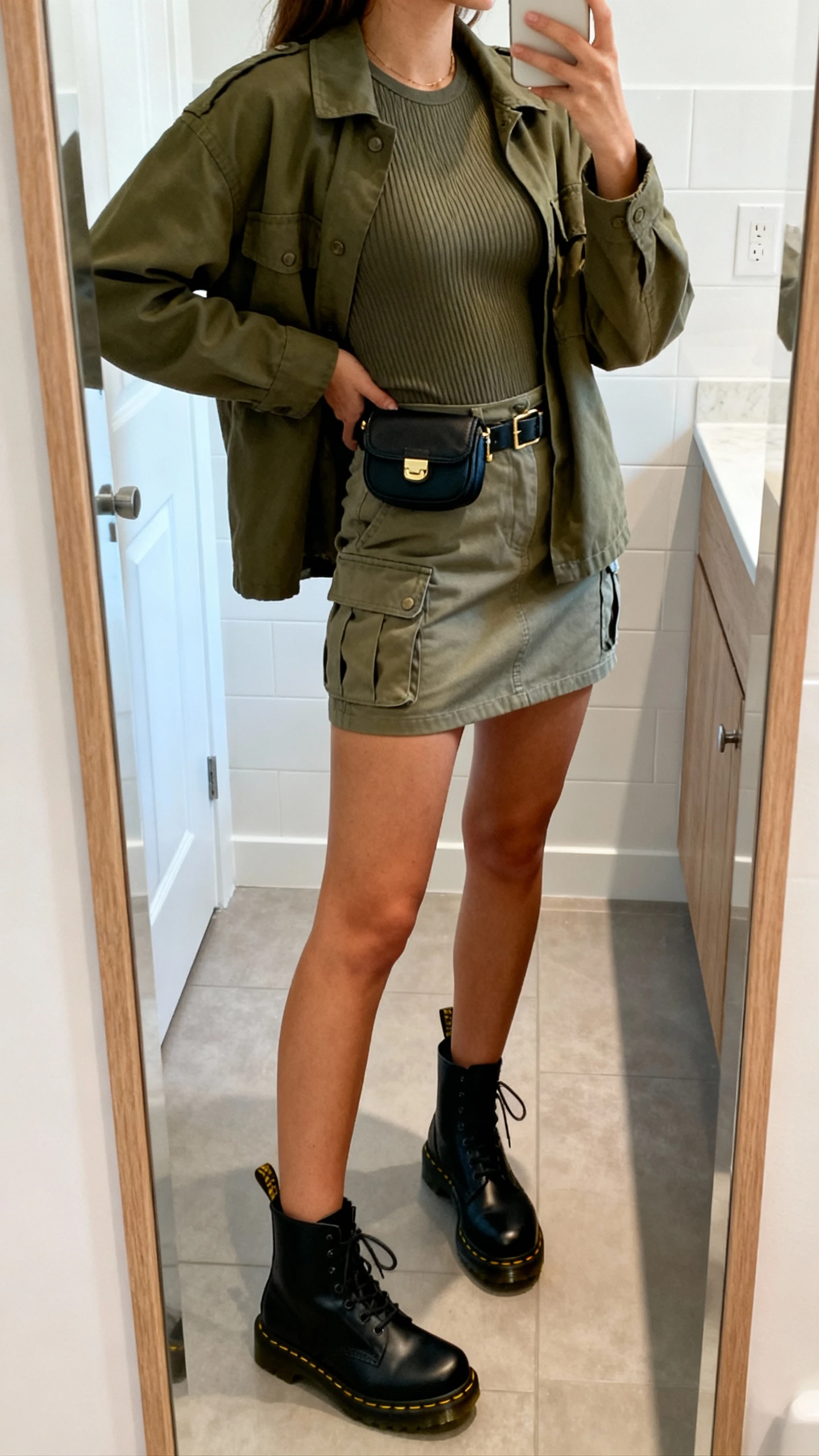 iPhone side-angle mirror selfie of a woman wearing a cargo mini skirt with a fitted ribbed top, olive utility jacket, black Doc Martens, and a small belt bag, face not visible, bathroom mirror, bright indoor lighting, iPhone photo quality.