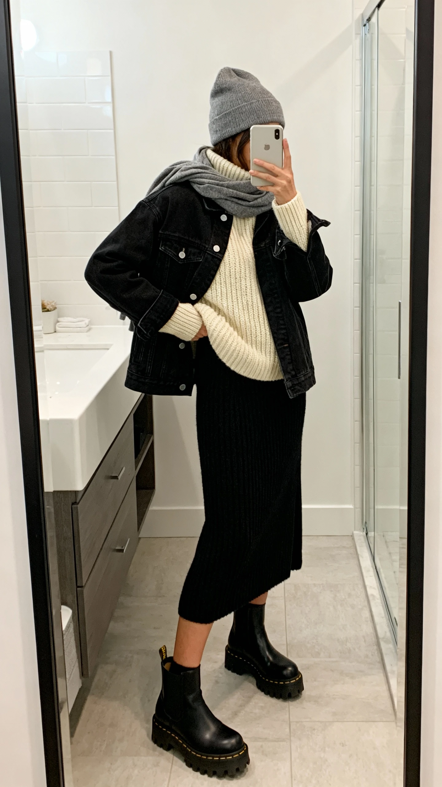 iPhone side-angle mirror selfie of a woman wearing a black denim jacket layered over a chunky cream turtleneck sweater, black knit midi skirt, black lug-sole Chelsea boots, and a gray beanie with a slouchy scarf, face not visible, bathroom mirror, soft indoor lighting, iPhone photo quality.