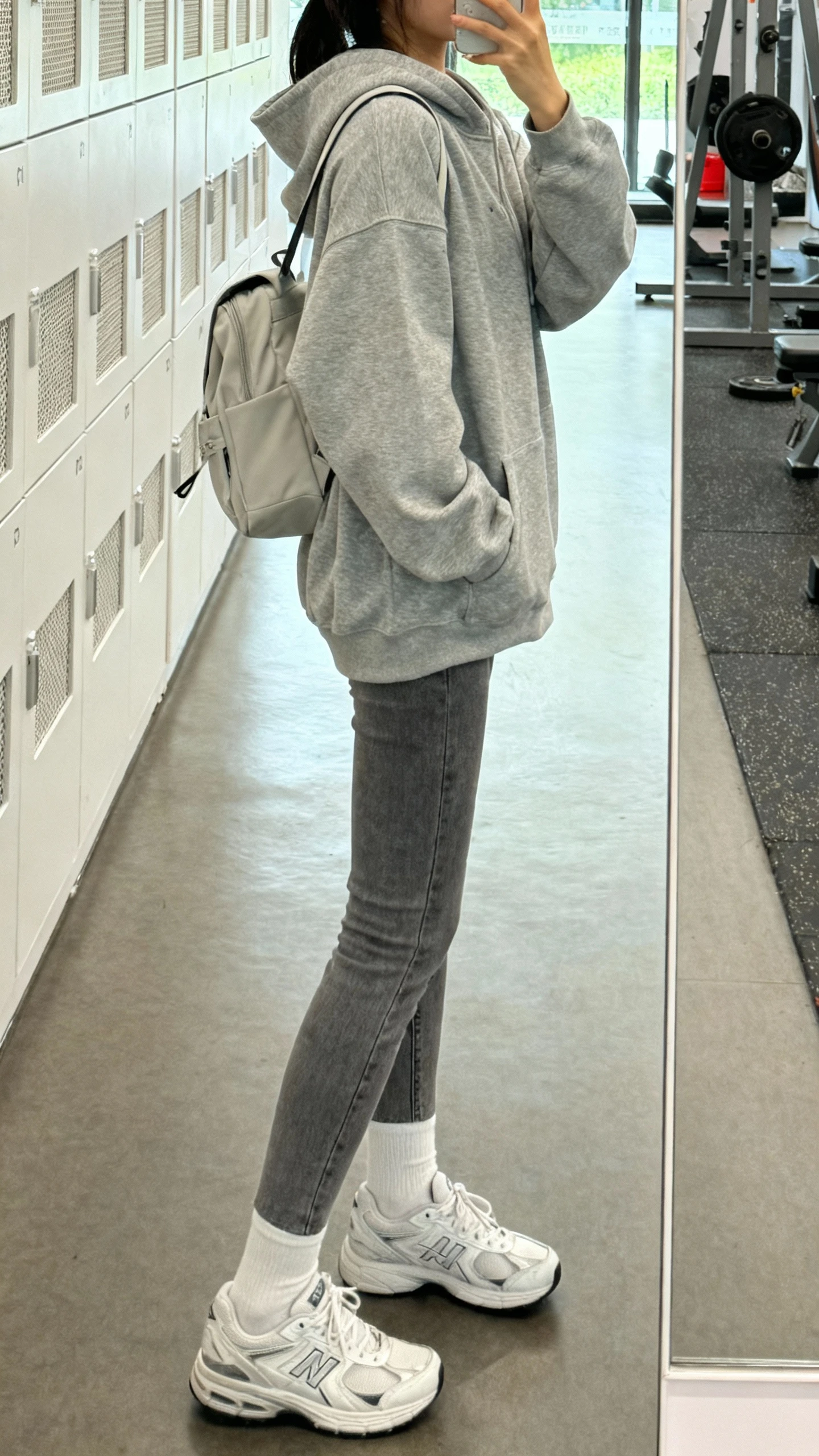 iPhone side-angle mirror selfie of a woman wearing grey jeans, an oversized heather grey hoodie, sleek white trainers, white ankle socks, minimal backpack, face not visible, gym locker room mirror, bright indoor light, iPhone photo quality.
